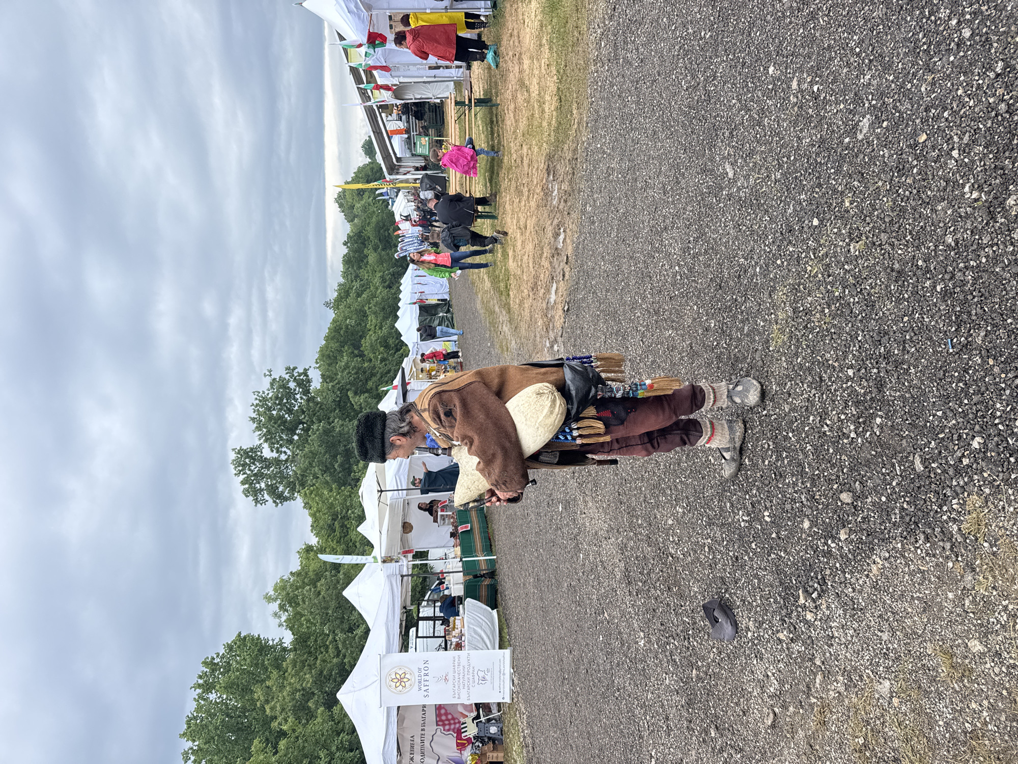 The image shows a person dressed in traditional clothing walking through an outdoor market or festival. The person is wearing a brown jacket, brown pants, a black furry hat, and striped socks with grey shoes. They are carrying a cream-colored pouch or bag and a bundle of items, possibly brushes or sticks.
The background features white tents or stalls set up in a row, suggesting a market or fair. People can be seen browsing and interacting with the stalls. The ground is covered in small stones or gravel. Trees are visible in the middle distance, and the sky is overcast with grey clouds.
Based on the text visible on one of the tents, the event is likely related to Bulgarian saffron products, with the text reading "WORLD OF SAFFRON" and including Bulgarian text as well. This suggests the location could be Sheremetia, Bulgaria, though the image doesn't provide enough detail to confirm the specific city.