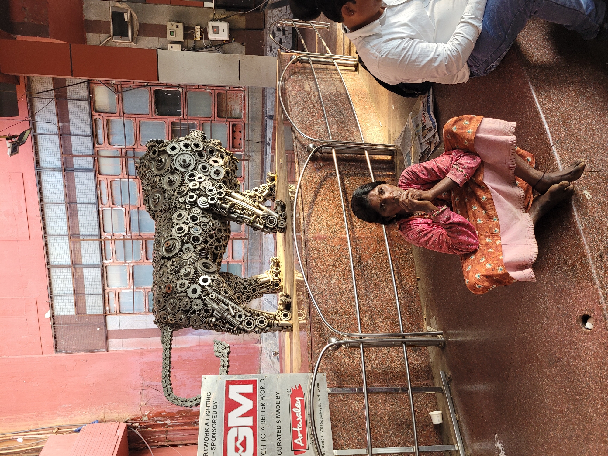 The image captures a scene in an urban environment with a juxtaposition of art and human presence. A sculpture of a tiger, constructed from gears and mechanical components, stands near a metal railing. Beside the tiger, a person is sitting on the floor, wearing a pink and orange patterned outfit. Behind the tiger is a building facade with square windows, and the background includes electrical wiring and components. A sign next to the tiger identifies the artwork as sponsored by "GM" and curated and made by "Artwaley." There is another person in the background sitting with their back turned to the camera and wearing a white shirt and jeans. The environment appears to be a walkway or public space with tiled flooring.