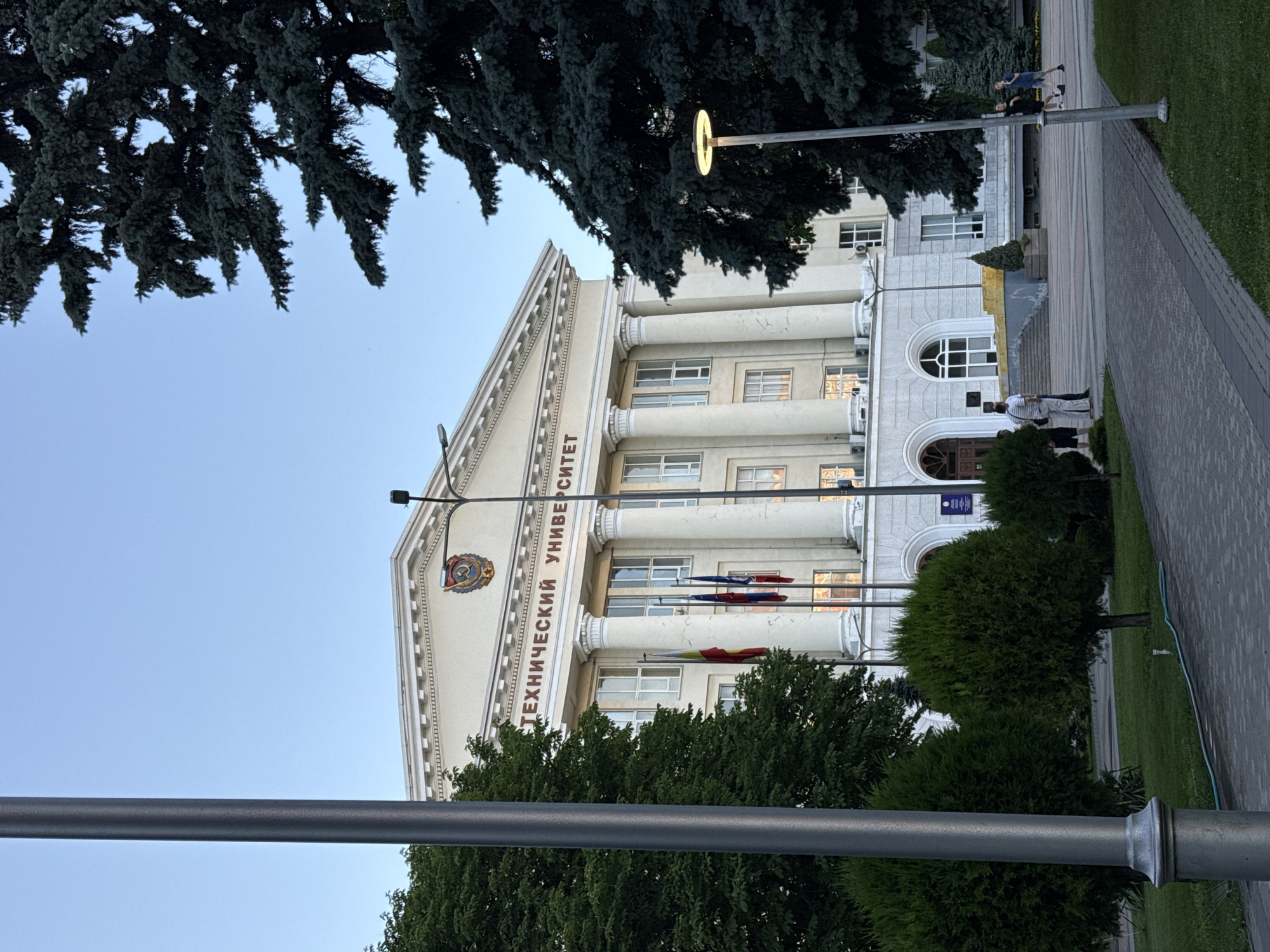 The image shows the Rostov State University of Economics building in Rostov-on-Don, Russia. The building is a large, classical structure with columns and an arched entrance. Text above the entrance reads "ТЕХНИЧЕСКИЙ УНИВЕРСИТЕТ," which translates to "Technical University." A coat of arms is visible above the text. Several flags are planted in front of the building. Trees and manicured bushes are present on the grounds, and a paved walkway leads towards the building. A streetlamp with a glowing light is visible to the right. Two individuals are walking on the walkway in the foreground, and two other individuals are standing near the entrance of the building on the left. The sky is clear and blue.