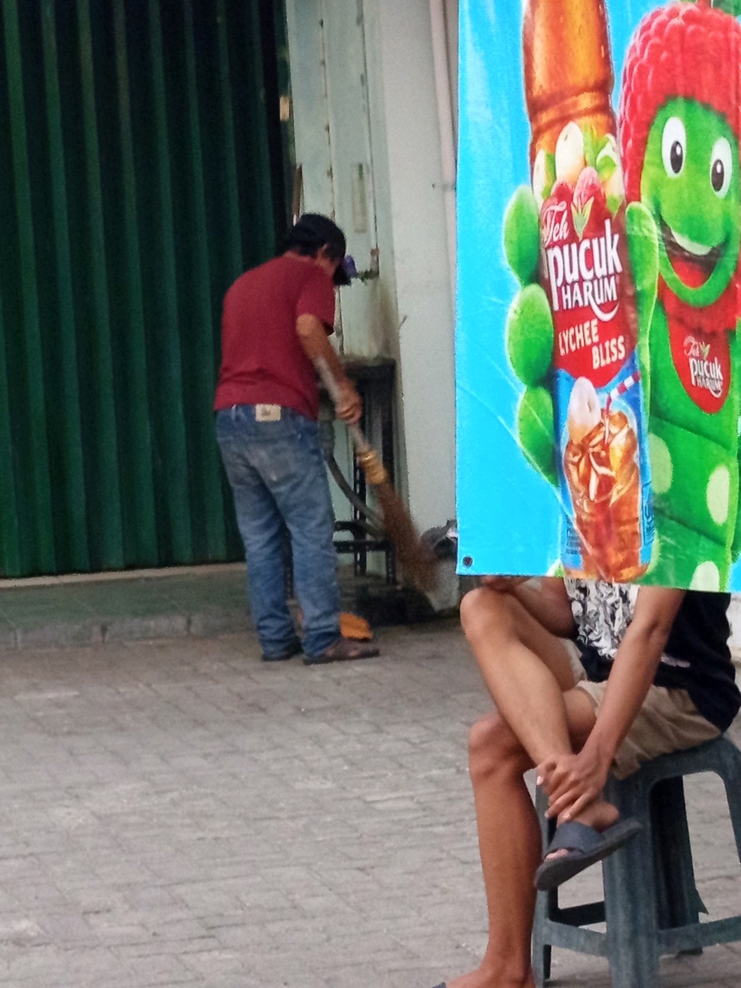 In a Karawang, Indonesia, outdoor scene, a man wearing a red t-shirt and blue jeans sweeps the paved ground with a broom. He is standing to the left of a bright blue banner displaying an advertisement for "Teh Pucuk Harum Lychee Bliss," which features a cartoon frog character and images of lychees and iced tea. To the right, another person, partially obscured by the banner, is seated on a green plastic stool, wearing a black t-shirt and beige shorts, with one leg crossed over the other and their hand resting on their knee. The environment appears to be an urban street or sidewalk, with a textured gray pavement and a dark green corrugated metal structure in the background.