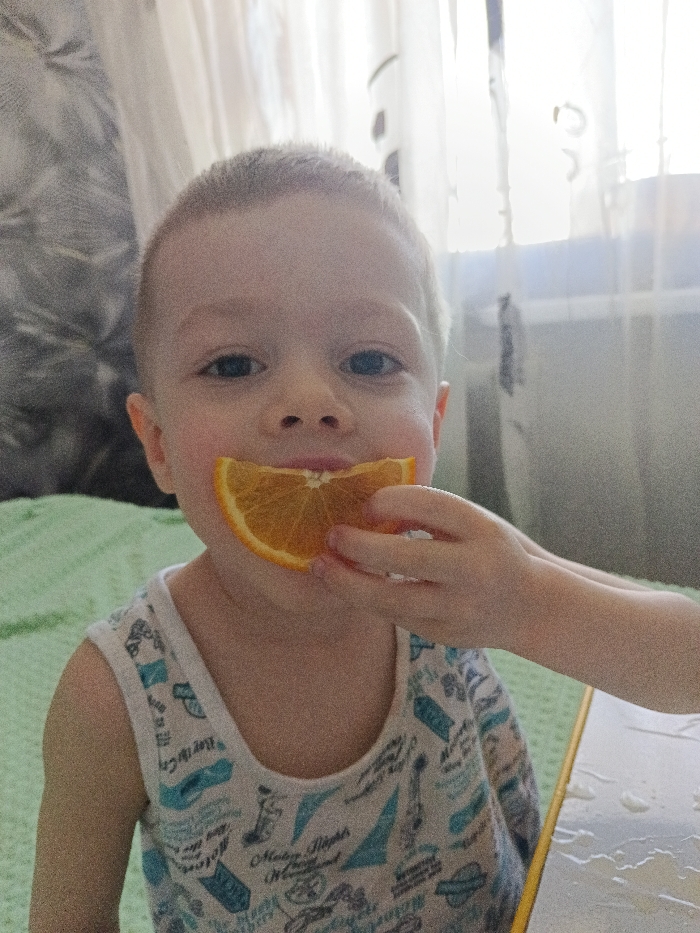 The scene is an interior domestic space within the city of Chaikovsky, Russia. A young Caucasian child, estimated to be between two and four years old, with short, light-colored hair and blue eyes, is depicted from the chest up, looking directly forward towards the camera with a slight smile. The child is wearing a white sleeveless undershirt or tank top with various blue and grey graphics, including text like "Mickey Mouse Wonderland" and "By the Sea." A wedge-shaped slice of an orange, approximately 5-7 cm in length, is held horizontally against the child's upper lip and chin. A light-skinned hand, visible from the wrist, holds the orange slice with the thumb and index finger, extending from the right side of the frame. Behind the child, to the left, is a textured green fabric, possibly a blanket or bedspread. Further to the left and upper left, a patterned grey and white wallpaper or wall covering is visible. Directly behind the child, a window covered by translucent white curtains is a source of bright ambient light, causing some overexposure in the background. In the lower right foreground, a light-colored surface, possibly a tabletop with a yellow edge, shows several small droplets of clear liquid. The lighting suggests a daytime setting.