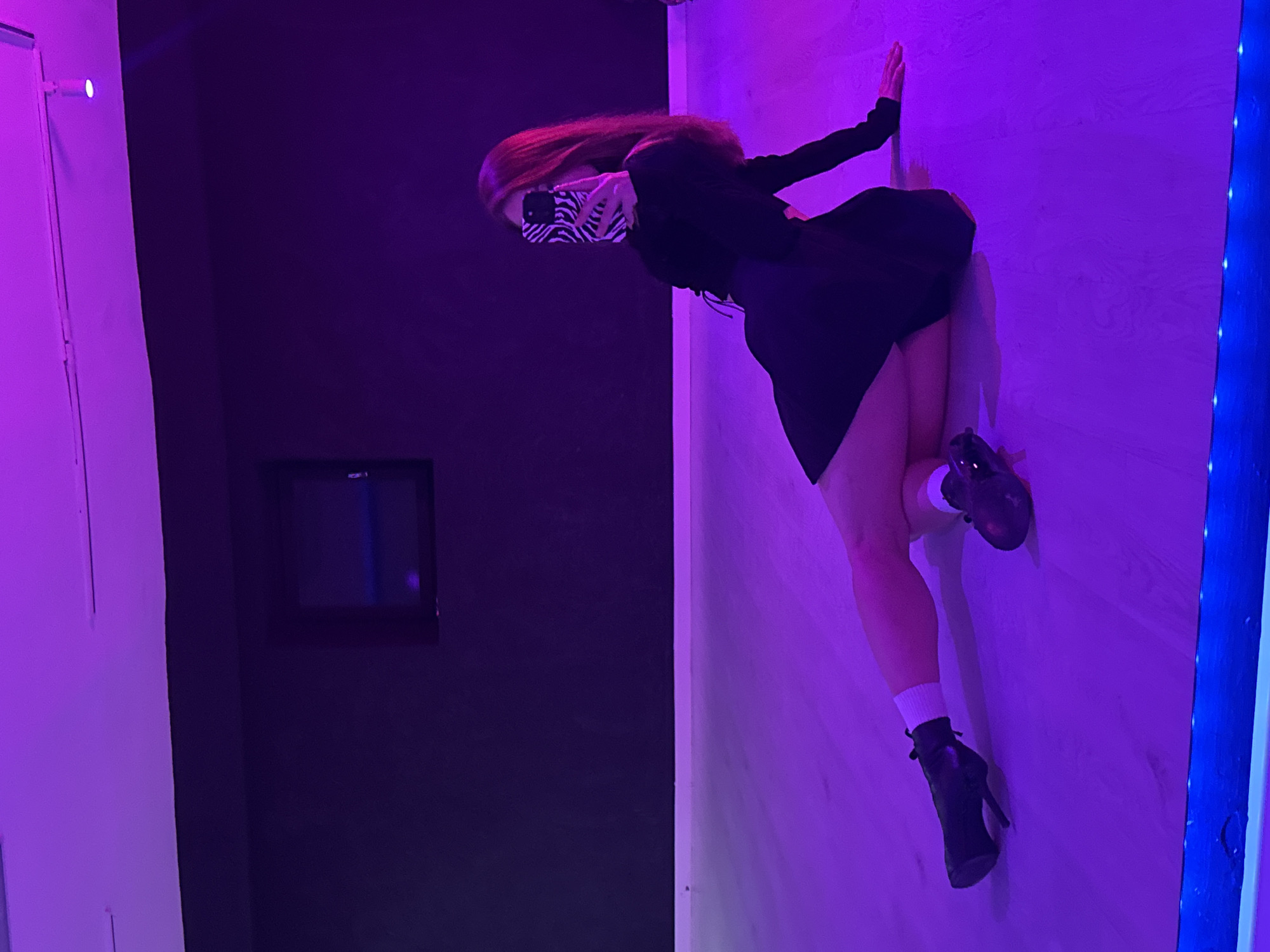 The image displays a person, likely female, creating an optical illusion within an indoor setting. The individual is oriented horizontally across the frame, appearing to stand on a light purple, possibly wooden-textured wall, as if defying gravity. One hand is pressed against this wall for balance, while the other holds a smartphone with a distinctive black and white zebra-print case, which partially obscures their face. Long, reddish-brown hair cascades "downwards" from their head, enhancing the illusion of an altered perspective.
The person is dressed in a black long-sleeved top, a black flared skirt, white ankle socks, and black high-heeled ankle boots.
The room is dramatically illuminated by artificial purple and blue lighting. The wall the person is "standing" on glows with a vibrant purple hue, while the background wall is a darker, deep purple or black. On this darker wall, a small, dark rectangular object, possibly a screen or monitor, is visible in the lower-left background, with blurry white text that might read "ROG_1" or "ROG-1" faintly discernible. A vertical strip of blue LED lights runs along the far right side of the image. The scene suggests an interactive art installation or a specially designed room for photography, aimed at creating an immersive, gravity-defying experience. The provided location context for this image is Chaikovsky, Russia.