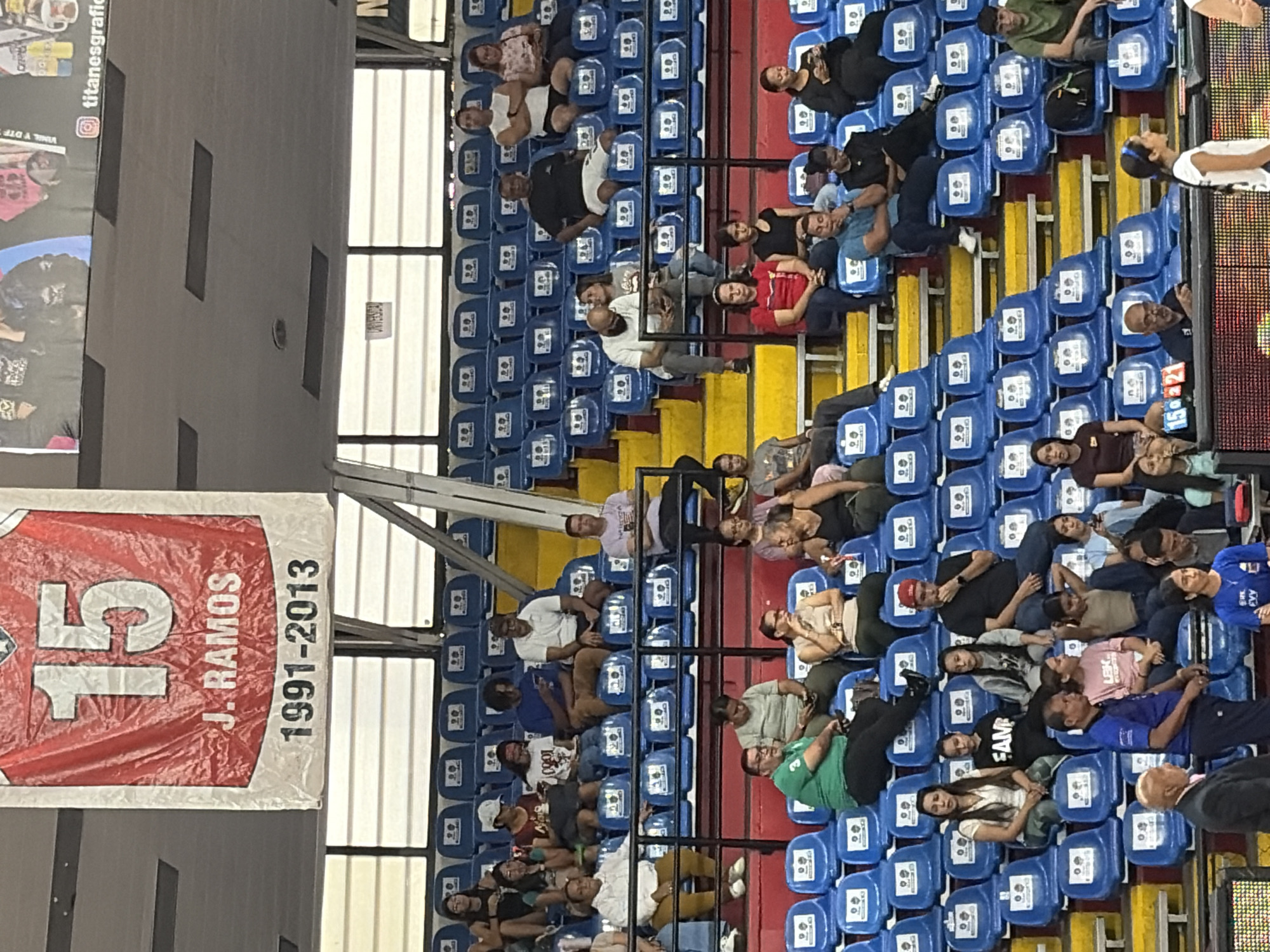 The image shows a partial view of an indoor stadium seating area with spectators occupying blue seats. There's a banner visible, red with white text: "15 J. RAMOS 1991-2013." Some spectators are sitting, some are reclining, and the view is from an elevated position facing the seating. Location details are not discernible in this image.