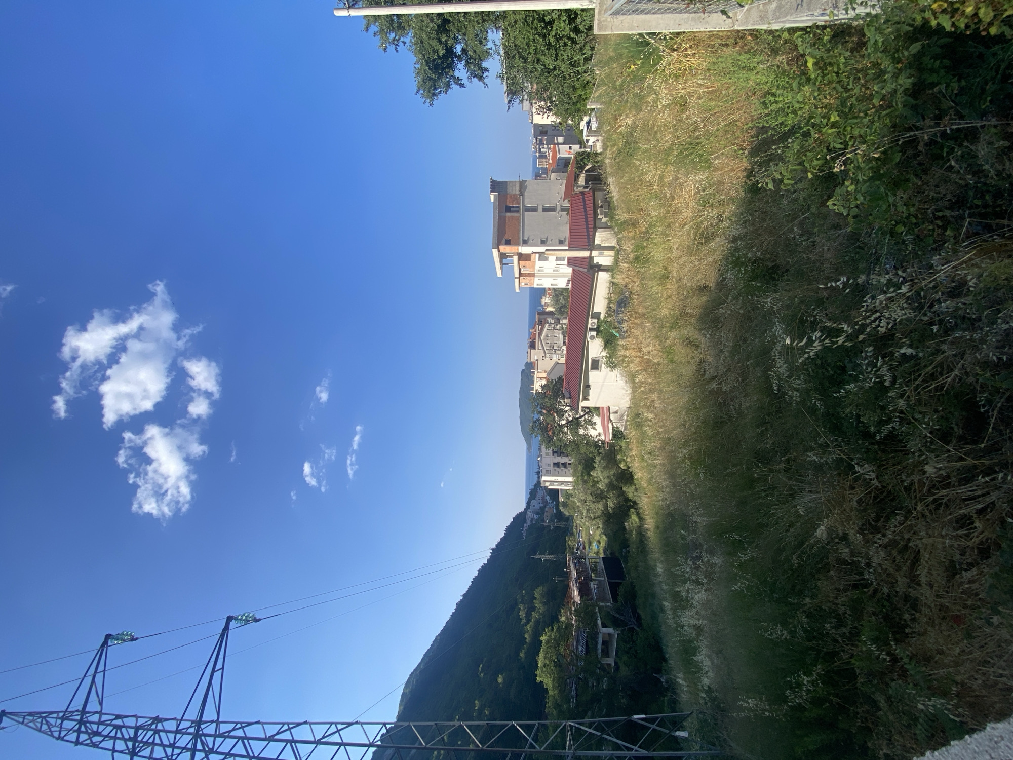 The image depicts a residential area in Budva, Montenegro, on a clear day with a bright blue sky and scattered white clouds. In the foreground, a tall, dark metal electricity pylon stands to the left, with power lines stretching across the frame. A slope covered in dry grass and green bushes dominates the mid-ground. Several buildings, including multi-story residential structures, are visible scattered amongst trees and vegetation. One building in the center-right features a light-colored facade with reddish-brown accents and a flat, unfinished rooftop. In the distance, the Adriatic Sea can be seen, with hilly terrain and more buildings along the coastline. The overall impression is of a partially developed or developing urban landscape nestled against a verdant, mountainous backdrop. No people or specific actions are discernible in the image.
