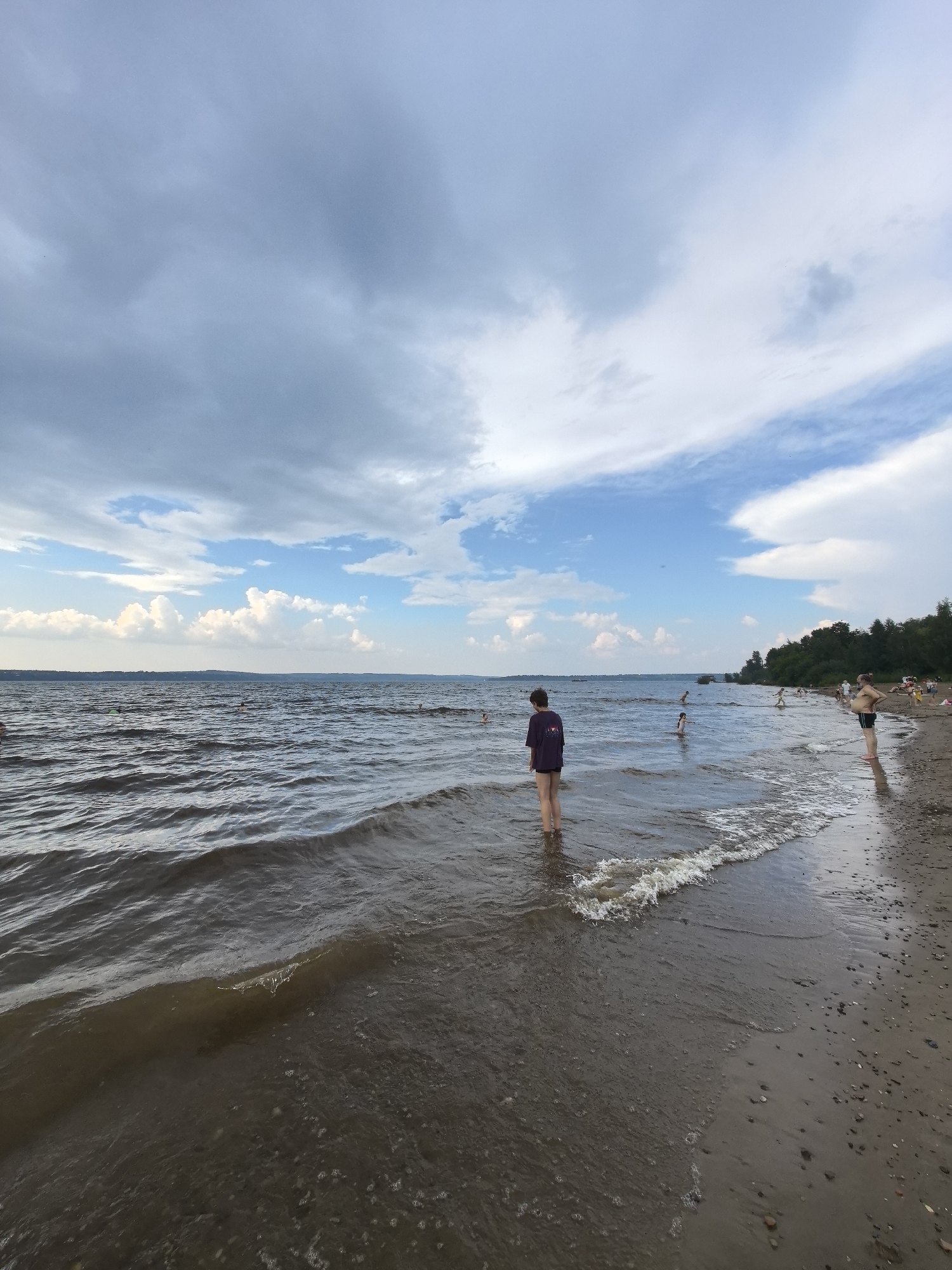 A wide shot of a beach scene on a cloudy day. In the foreground, a person stands partially in the water wearing a dark purple shirt and dark shorts. The water is wavy and brown, with small waves lapping at the shore. The beach is sandy with small pebbles. In the middle ground, several other people are in the water, some appearing to be swimming or wading. Further back, along the treeline, more people can be seen on the beach. The background shows a wide body of water with a distant treeline and a cloudy sky. The image appears to be taken at a recreational beach, possibly on the Kama River in Perm, Russia.