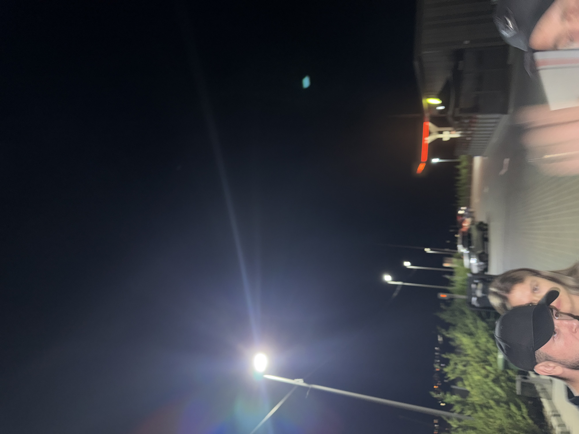 At night, a group of people are standing near a street lamp in Băcioi, Moldova. A man in a black baseball cap is in the foreground, with a woman next to him. In the background, there is a gas station with red and green lights. There are also several cars and trees visible. The sky is dark and clear, with a few stars visible.