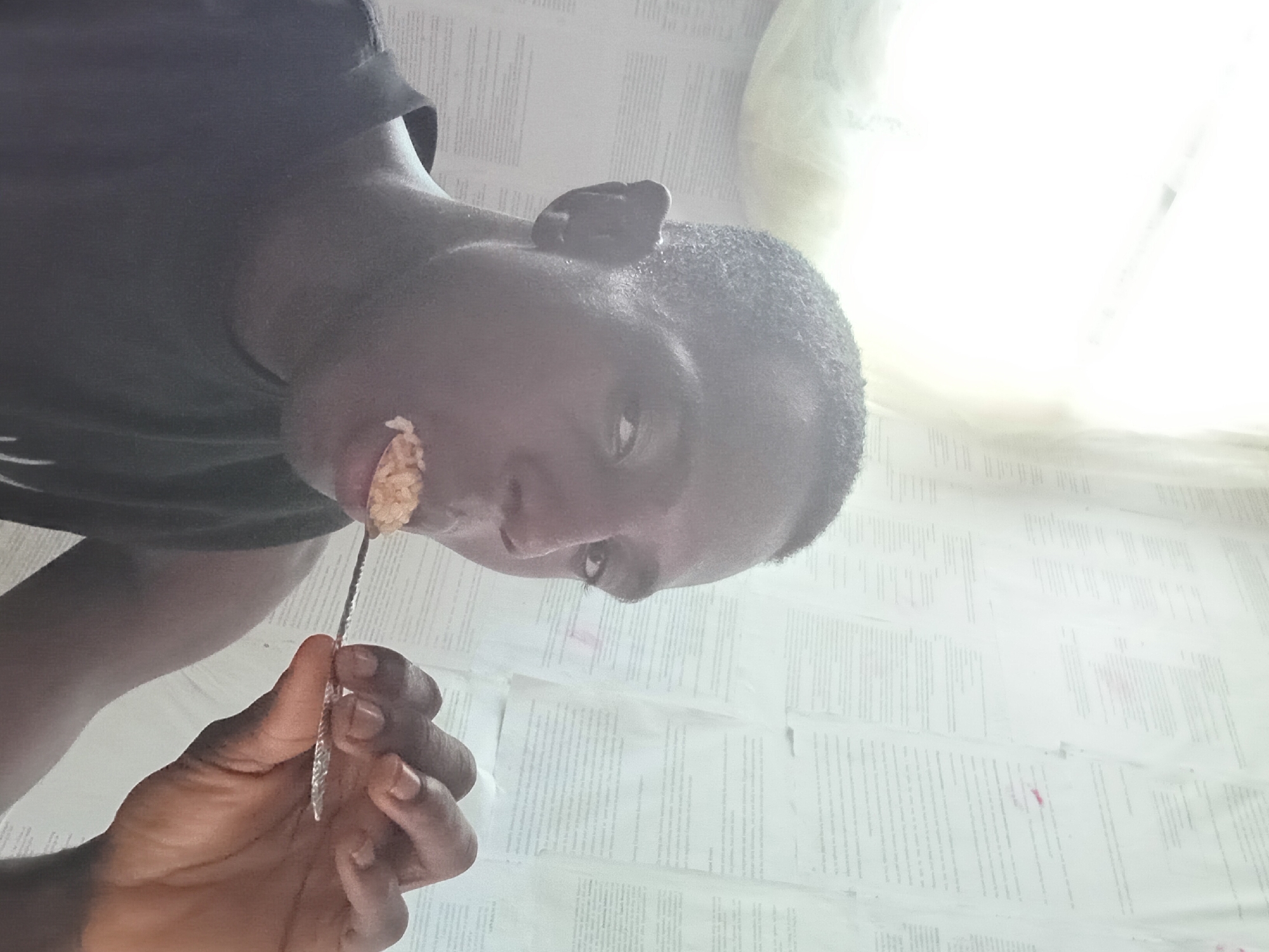 A person is holding a spoonful of food up to their mouth. The person is dark-skinned and wearing a dark t-shirt. The person is holding a spoon in their right hand. The spoon is holding a bite of food. The background appears to be a cluttered white surface with writing. The location is in the city of Akwete, Nigeria.