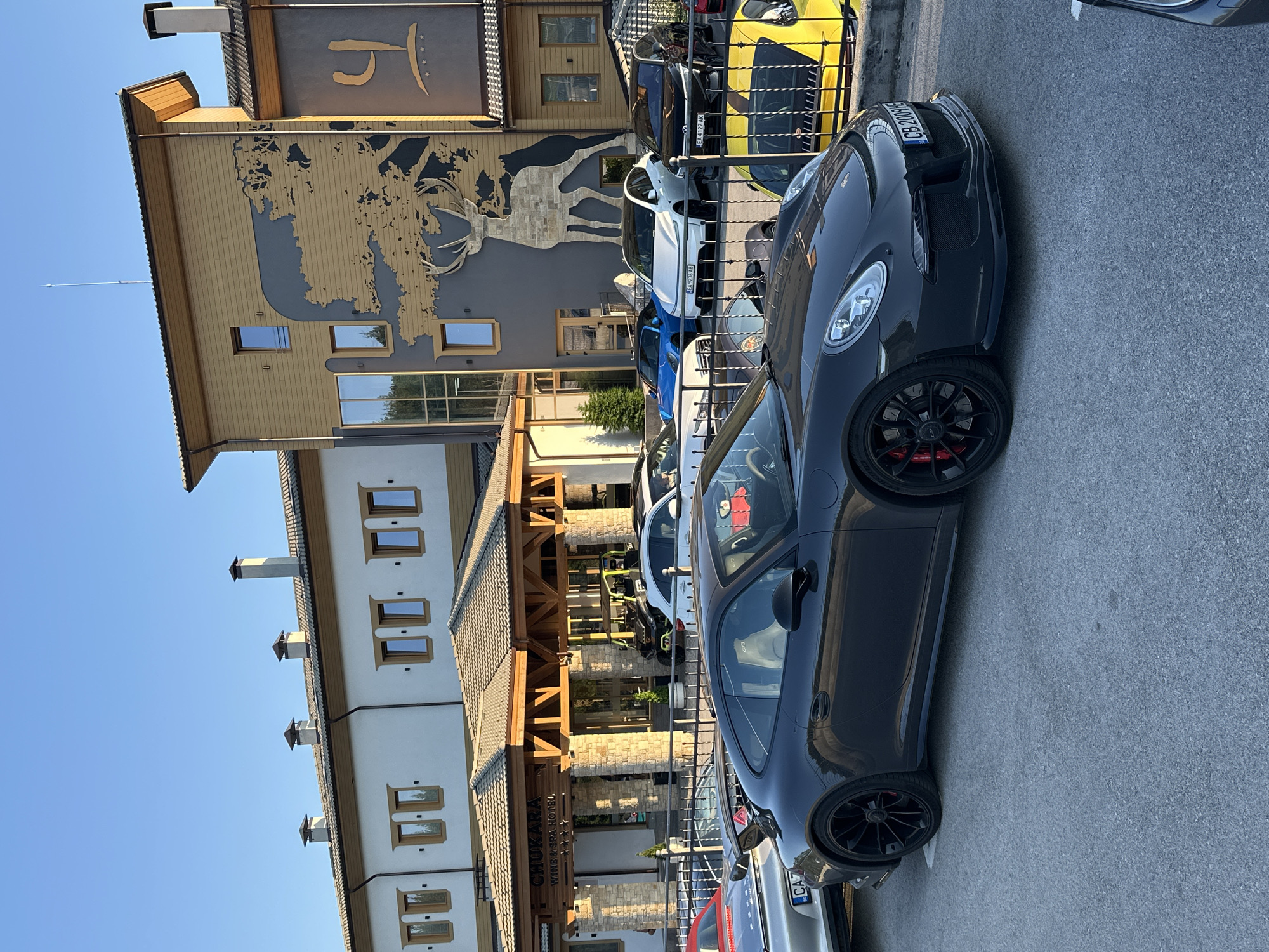 This media file captures a scene in Kerenite, Bulgaria, featuring several parked luxury cars, including a dark grey Porsche, a white Porsche, a blue sports car, and a yellow sports car. The cars are parked in front of a modern building with a distinctive architectural style, featuring a wooden facade and stone accents. The building appears to be a hotel or resort, with the text "CHUKARA WINE & SPA HOTEL" visible on a wooden sign.
In the background, a larger building with a tan and dark grey facade displays a large, stylized silhouette of a deer. The sky is clear and blue, indicating a bright, sunny day, likely in the late afternoon or early evening due to the low angle of the sunlight casting shadows. The overall atmosphere suggests a high-end establishment catering to car enthusiasts or those seeking a luxurious getaway. The presence of multiple high-performance vehicles hints at a gathering or an event related to these cars.