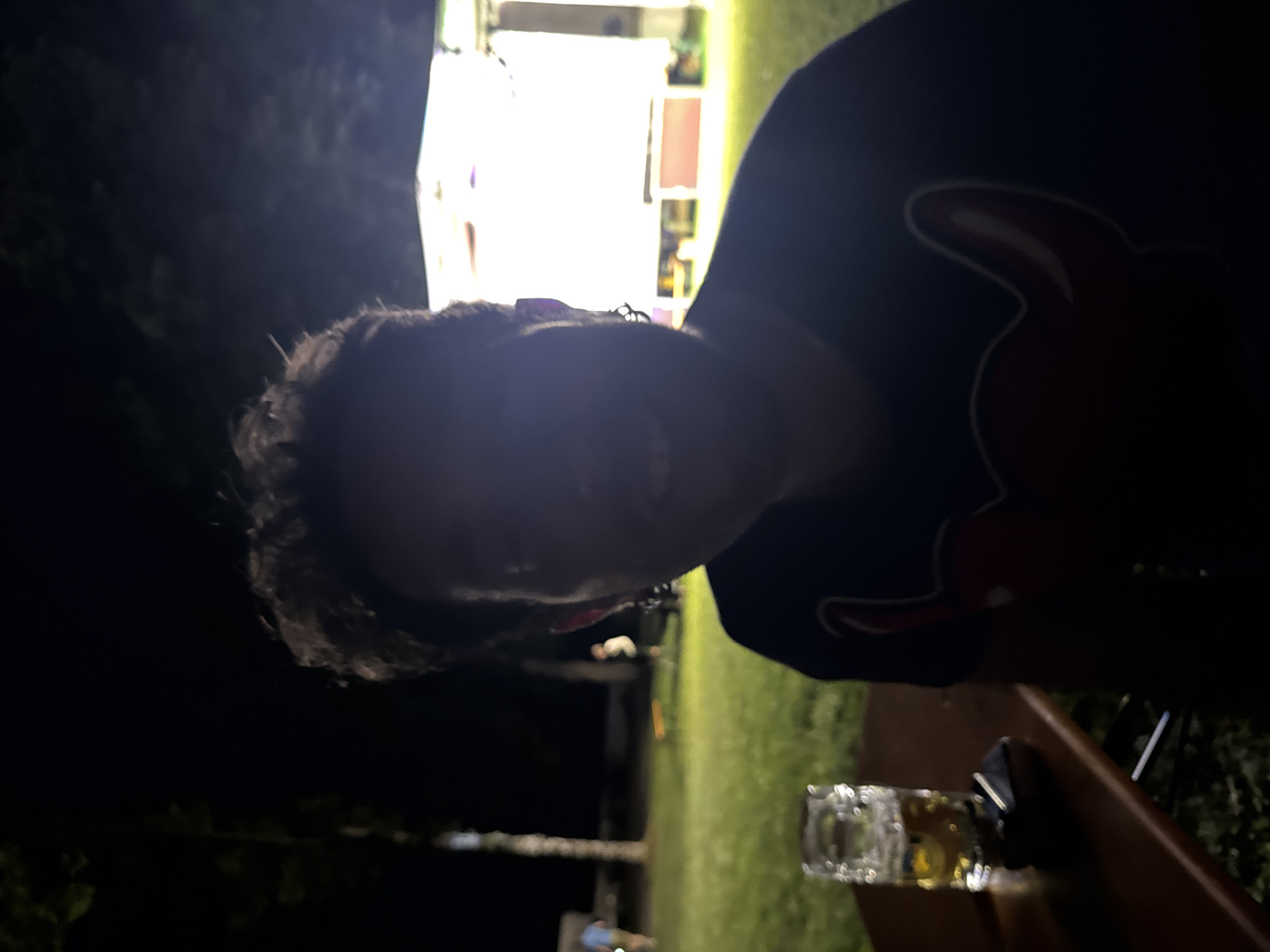 The image shows a person sitting at a wooden table outdoors at night. In front of the person is a glass of beer and what appears to be a small electronic device. The person is wearing a dark t-shirt with a red design on it. In the background, there is a brightly lit structure that resembles a stage or tent, and other people are visible in the distance, along with trees and grass. The setting suggests a festival or outdoor event taking place in Höhenkirchen-Siegertsbrunn, Germany. The lighting is low, with the main light source appearing to be artificial lights from the event. The focus is on the person in the foreground, though their face is partially obscured by shadow.