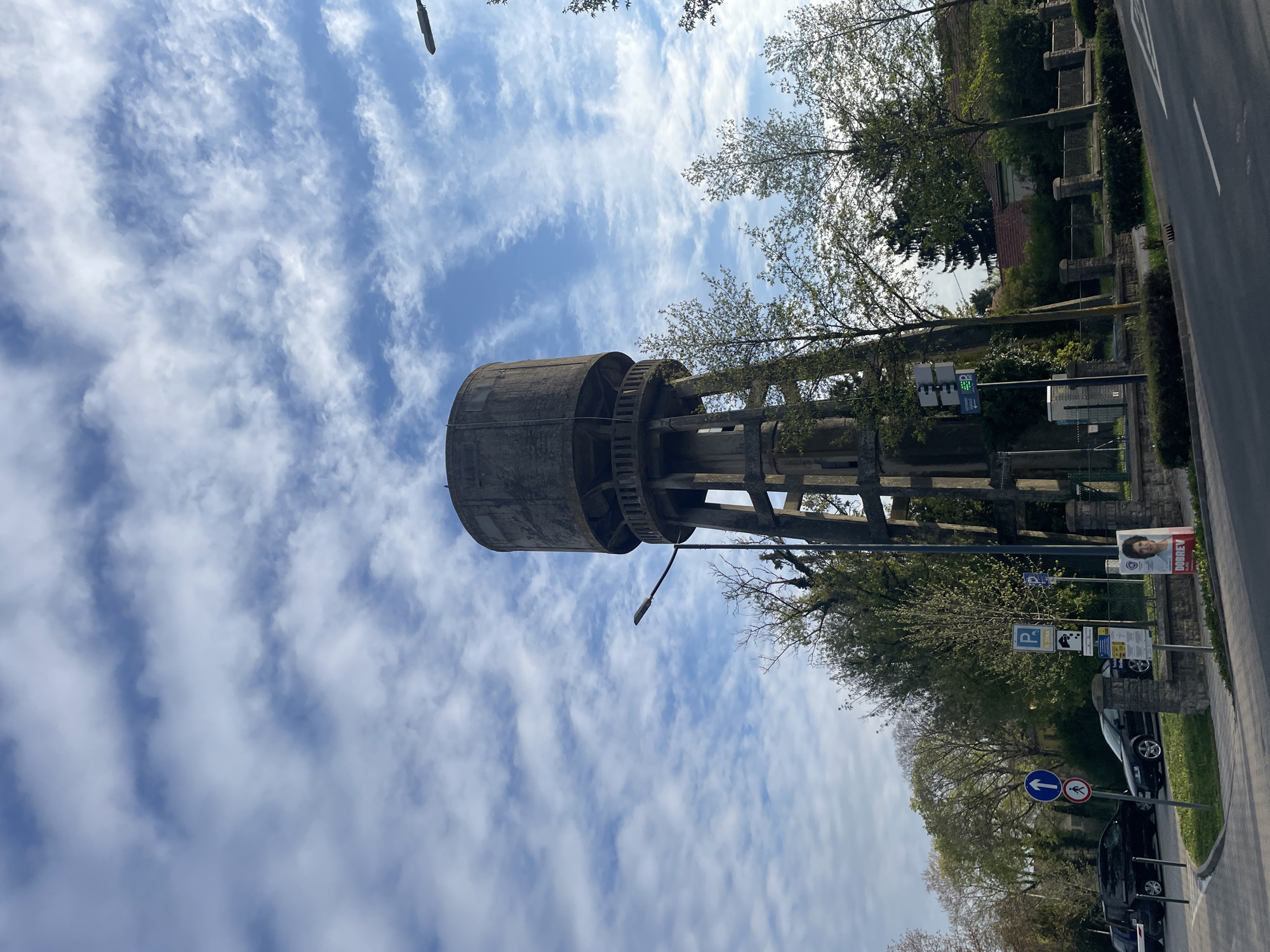 The image shows the water tower in Hévíz, Hungary, taken from a low angle. The cylindrical concrete water tank is elevated by a concrete framework. A street light is attached to the tower structure. The sky behind the tower is blue with many white clouds. To the right of the tower, there are trees, signs indicating parking, and a car parked along the side of the road. The road itself is asphalt. The overall scene is sunny and bright.