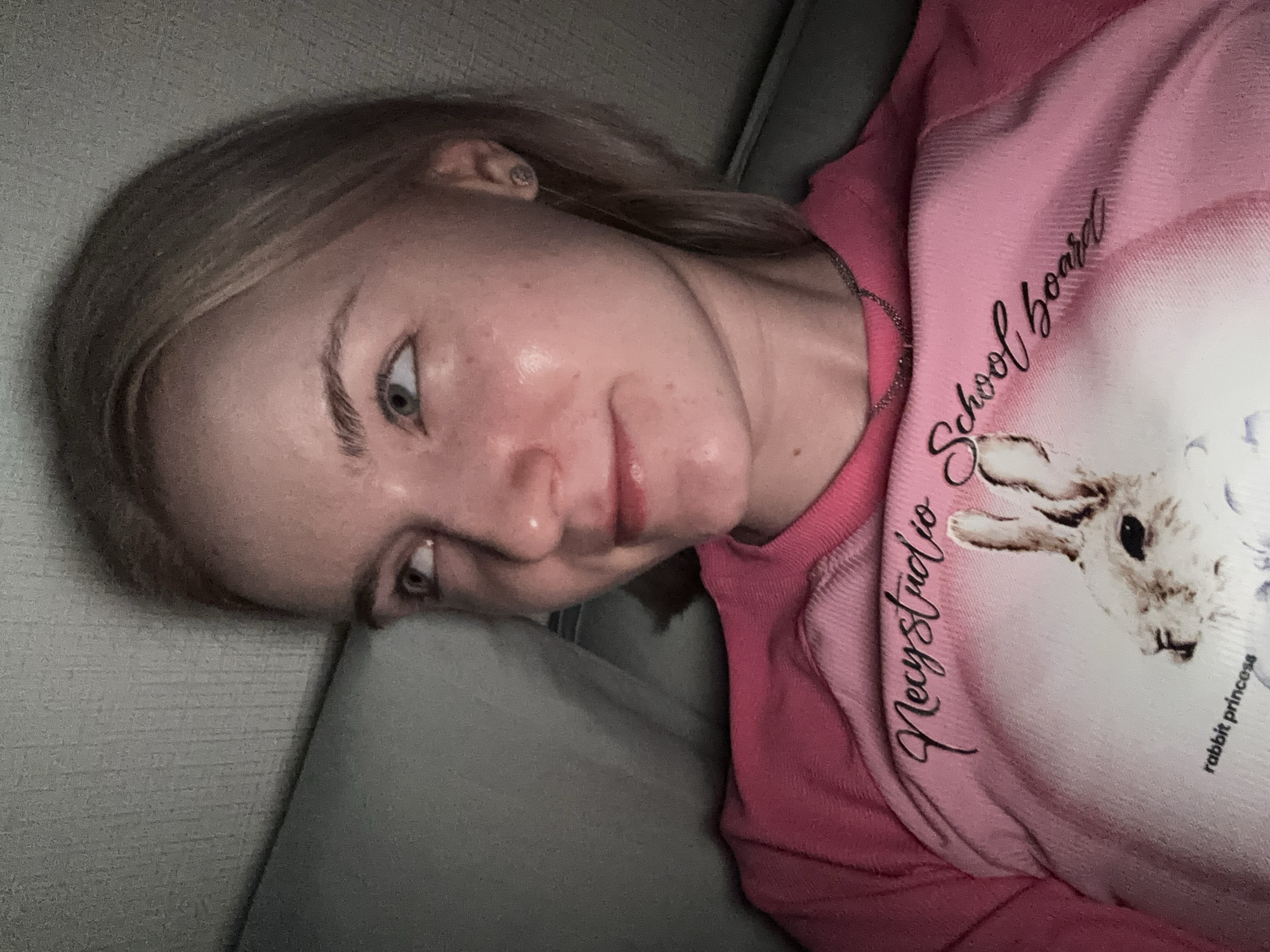 A close-up, angled portrait shot captures a young person, likely female, with fair skin and light-colored hair, lying down indoors. Her head is tilted to the side, and she gazes directly into the camera with a subtle, somewhat pensive or curious expression.
She is wearing a vibrant pink long-sleeved top. Prominently displayed on the top are several lines of text: "Mecystudio" and "School board" in a stylized script font, and "rabbit princess" in a smaller, standard font. A graphic of a rabbit is partially visible on the shirt. A thin silver chain necklace is worn, and a small earring is visible in her right ear.
The background suggests an indoor setting, possibly on a sofa or bed, with soft, grey-toned upholstered textures and a textured wall. The lighting is dim, indicating an evening or nighttime scene. The person's skin exhibits a natural sheen, possibly due to the ambient light. This scene is located in Vsevolozhsk, Russia.
