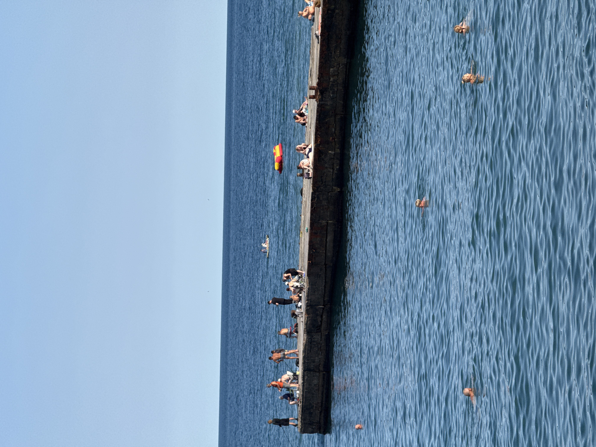 The image depicts a concrete pier extending into the blue sea under a clear sky. Several people are present on the pier, with some fishing, others sunbathing, and some appear to be socializing. The sea has several individuals swimming or wading. In the distance, a red and yellow inflatable tube is visible on the water, and another person is on a paddleboard. The pier is old and weathered. This scene is likely located in Partenit, Ukraine, a coastal resort town.