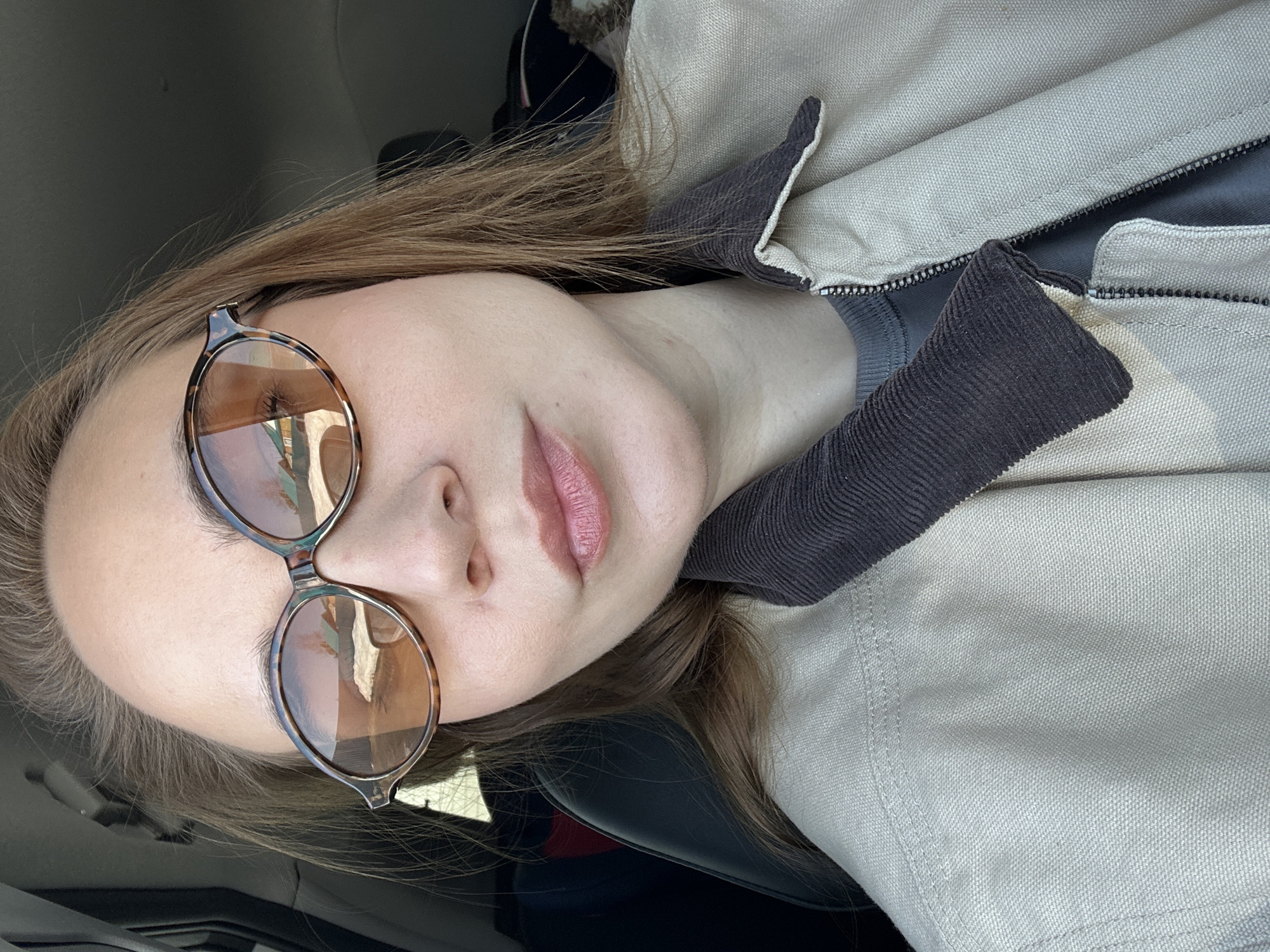 The image depicts an adult female subject seated within a vehicle, capturing a close-up selfie. The location is specified as the city of Kharnavy, Russia. The subject has fair skin, light brown hair, and is wearing tortoise-shell framed sunglasses with amber-tinted lenses. Reflections within the lenses show an outdoor environment featuring a structure with a light-colored roof and what appears to be ground covered in snow or a light substance. Her lips are lightly colored with pink lipstick and are slightly parted. She wears a light beige canvas jacket with a dark brown corduroy collar, partially unzipped to reveal a dark grey t-shirt underneath. The immediate background consists of the dark interior of a vehicle, including a dark car seat behind her left shoulder and a dark headliner above. The subject is looking directly at the camera with a neutral expression.