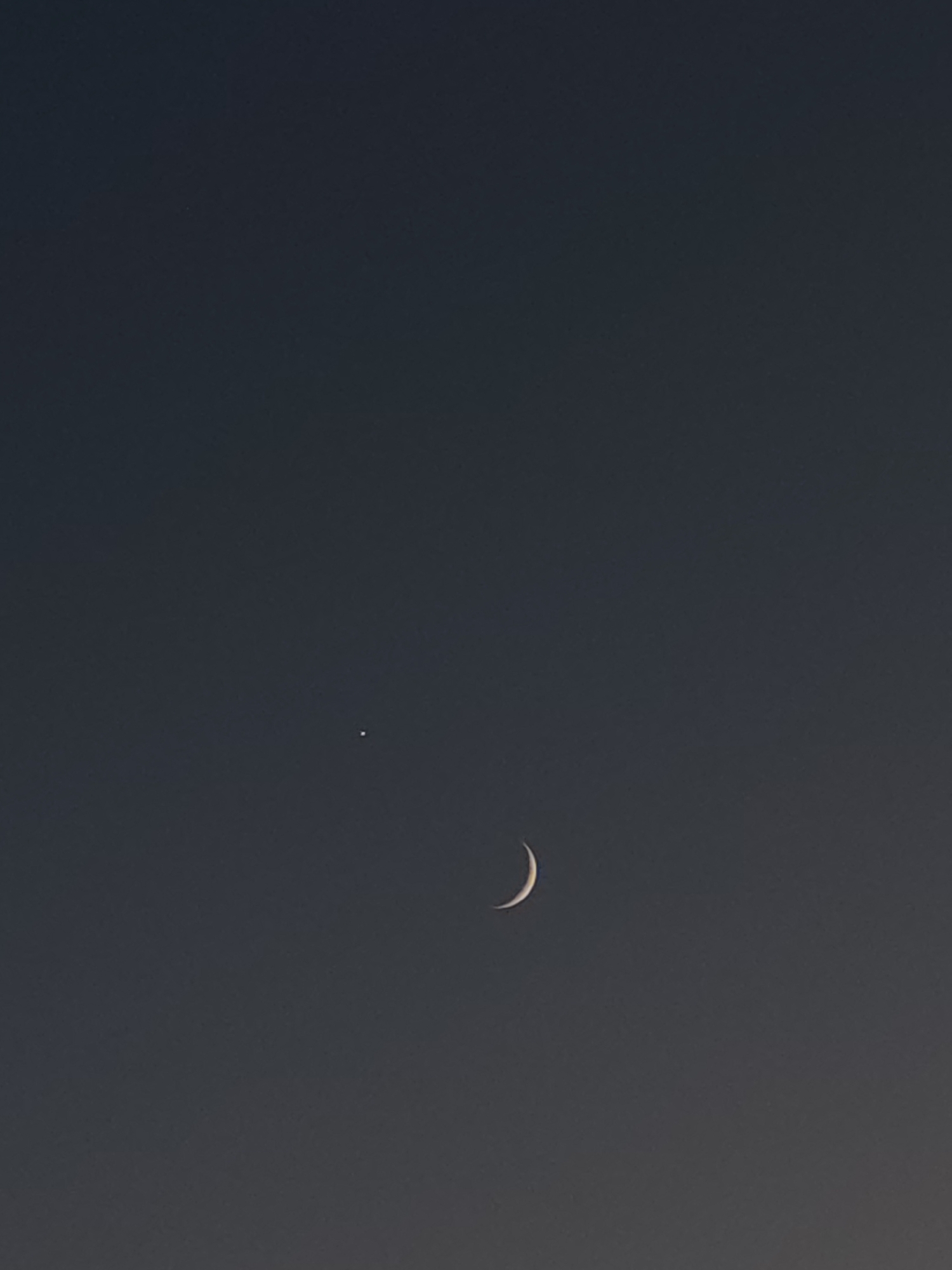 An image shows a dark sky with a crescent moon and a bright point of light. The crescent moon is visible in the lower right portion of the frame. A single bright point of light, likely a planet or a star, is located to the left and slightly above the moon. The background is a deep, uniform dark gray, suggesting it is nighttime or twilight. There are no other discernible objects, people, or environmental features visible in the image. The location is unknown.