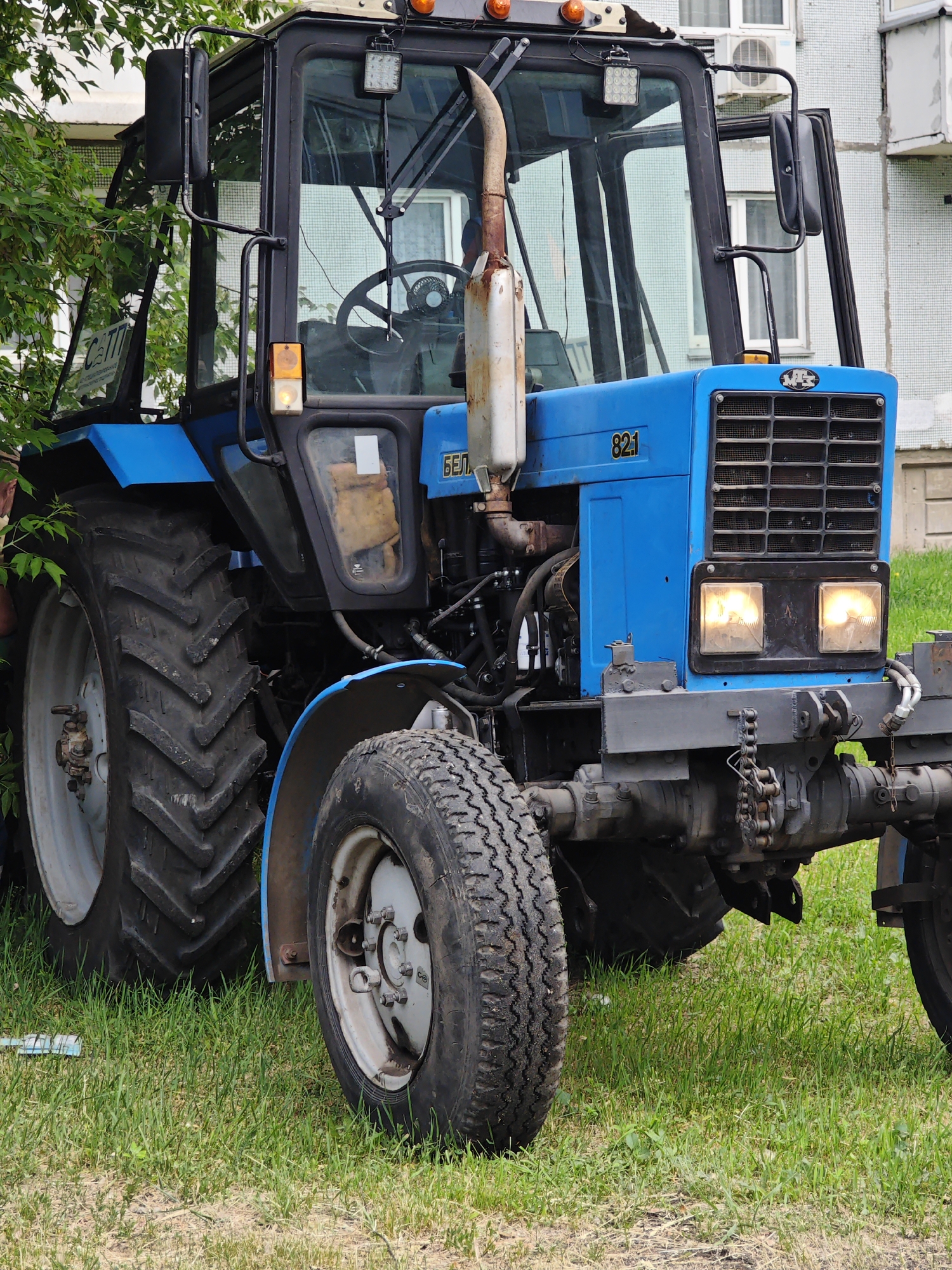A blue Belarus tractor, model 821, is parked outdoors on a grassy area. The tractor has large, deep-treaded rear tires and smaller front tires. It features a black cab with large windows, a visible steering wheel, and an orange turn signal. The front of the tractor has two illuminated headlights and a grille. A rusty exhaust pipe extends vertically from the engine compartment. The background shows a building with multiple windows and an air conditioning unit, suggesting an urban or suburban setting. Green foliage from a tree is visible on the left side of the image. The image was taken in the city of Krasnoyarsk, Russia. There are no people or any discernible actions or interactions depicted.