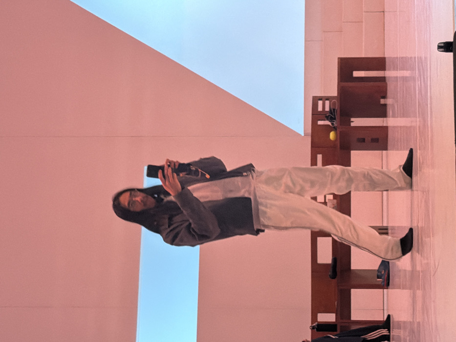 An indoor scene features a male individual with long dark hair and glasses, positioned centrally and looking forward. He wears a dark grey collared jacket over a light-colored top, white trousers with a dark stripe along the outer seam, and black socks, appearing shoeless. Both hands grasp a black smartphone, held upright as if recording or photographing. A small, wired microphone or earpiece is clipped to his top.
Behind the individual, a series of dark wooden, low-profile geometric furniture pieces are arranged. On one of these, a yellow spherical object and some black cables or accessories are visible. A dark blue shoe rests on the reflective floor near the furniture. To the far left, the lower legs of another person are partially visible, clad in dark pants with white stripes and dark footwear, alongside a black water bottle.
The background consists of a plain wall, illuminated with a warm, pale pink light. Two prominent, cool light-blue shapes are projected or illuminated onto this wall: a horizontal rectangle on the left and a large triangle on the right. The floor is a highly reflective surface, mirroring elements of the scene. The precise location within Paris, France, cannot be determined from the visual information presented.