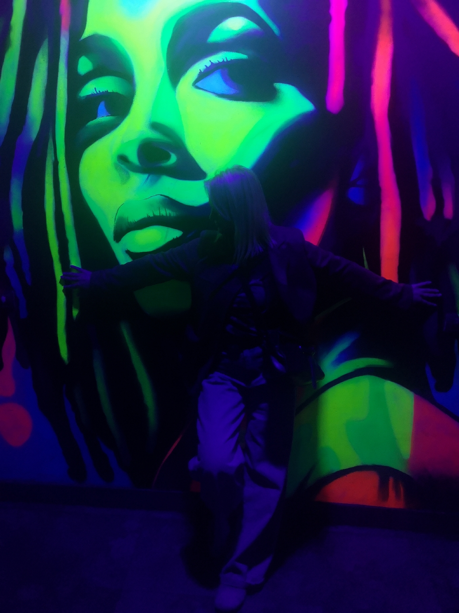 The image shows a person standing in front of a large, neon-lit mural of a woman's face with dreadlocks. The mural is predominantly green, with highlights in pink, blue, and red. The person in the foreground has their arms outstretched, and they are wearing a dark jacket and light-colored pants. The room is dimly lit with a blue hue. The location is Chaikovsky, Russia.