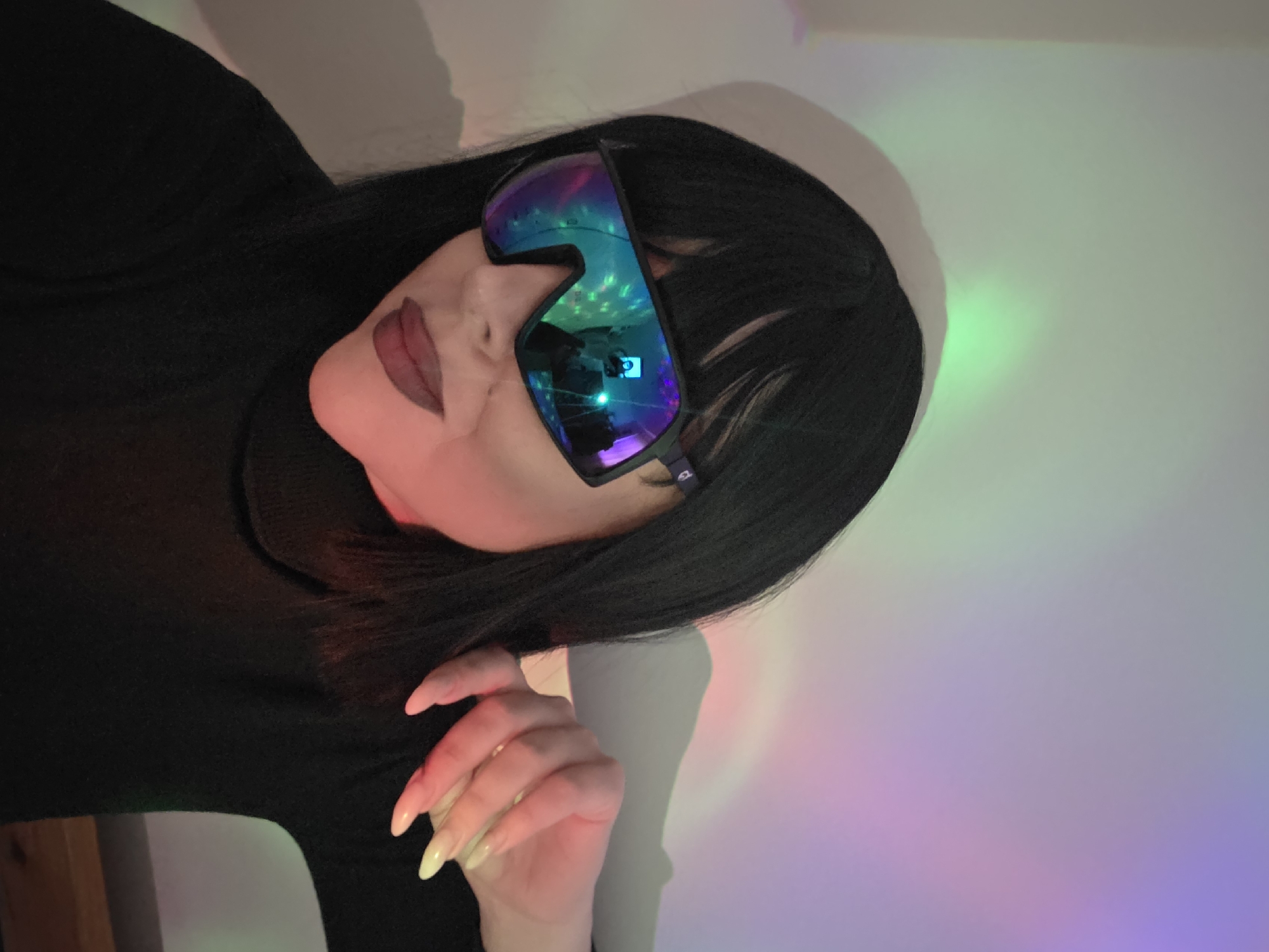 A woman with dark hair and lipstick is wearing sunglasses that reflect colorful lights. She is wearing a black turtleneck shirt and has long, pointed nails. The background is a wall with a mix of green, pink, purple, and blue light shining on it. The location is unknown.