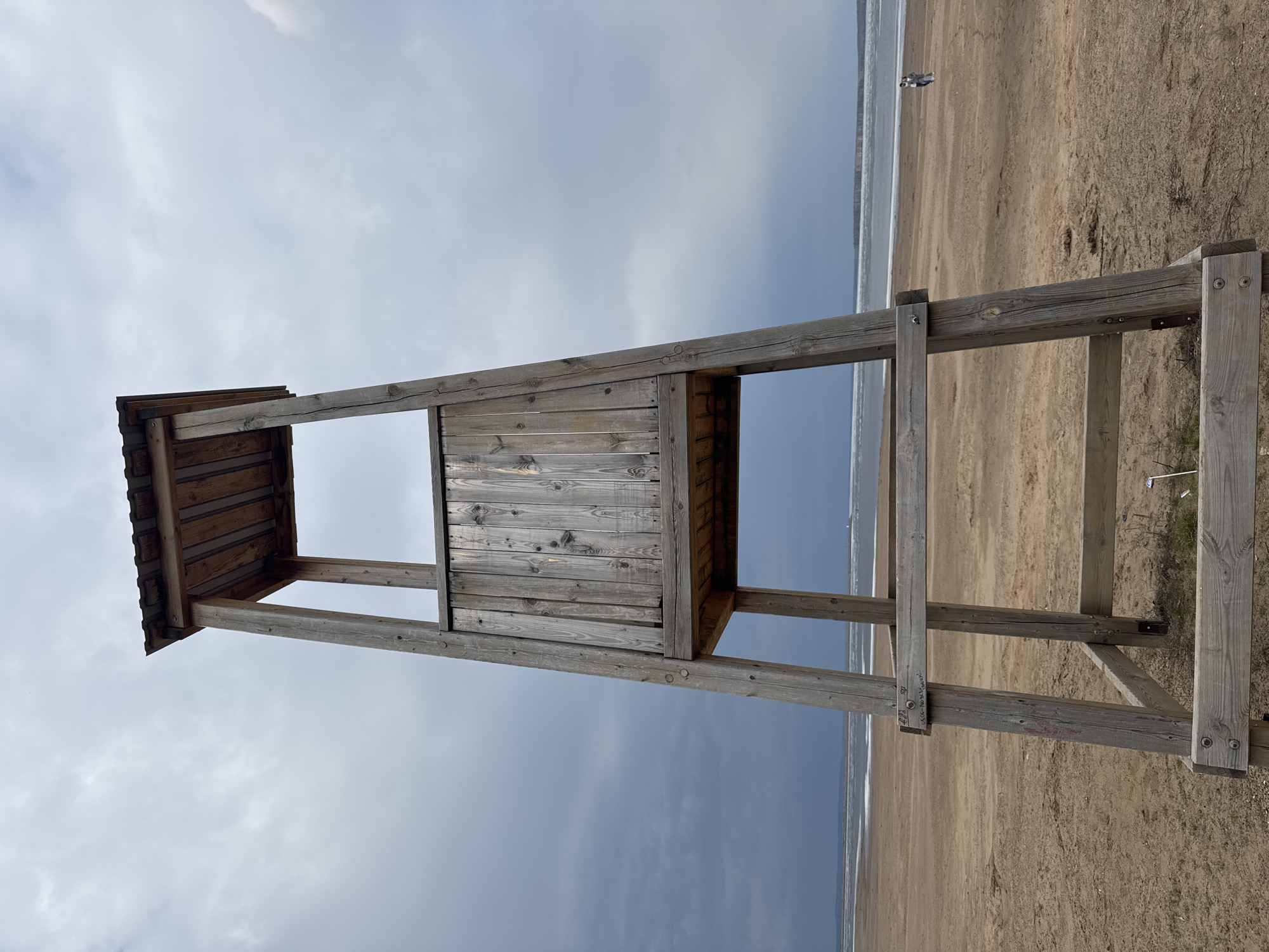 This image shows a lifeguard tower made of weathered wood standing on a beach. The tower has a small, enclosed platform with a roof. The beach is sandy and stretches out to meet the water, which is visible in the background. Two people can be seen walking in the distance along the shoreline. The sky is overcast with gray clouds. The location is likely Chaikovsky, Russia, given the prompt's parameters, although this cannot be definitively confirmed from the visual data alone.