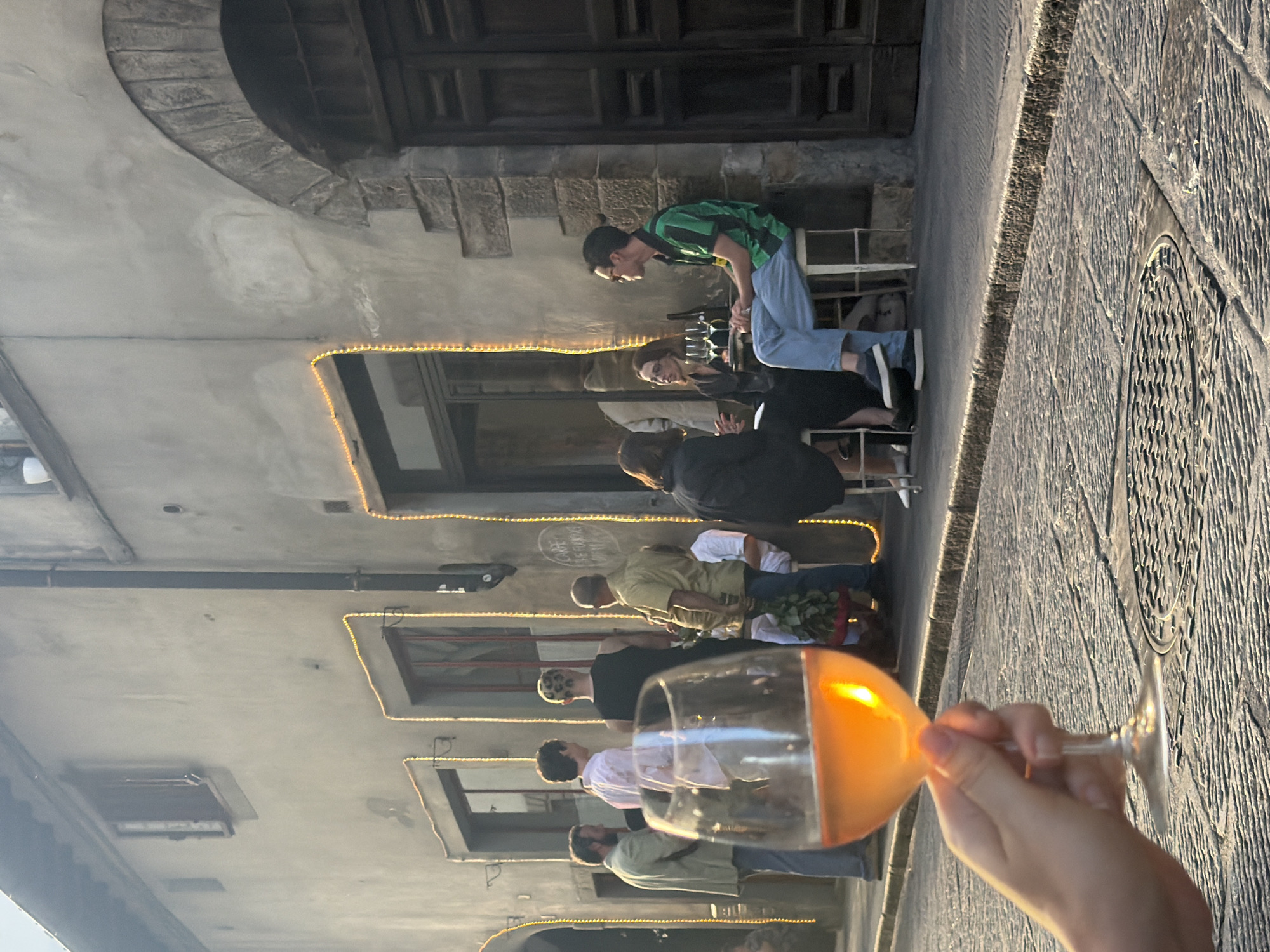 A group of people are gathered outside a building with an old stone facade, decorated with string lights. In the foreground, a hand holds a glass of rosé wine, with the glass obscuring a portion of the scene. The setting appears to be a street in Florence, Italy, with cobblestones visible in the foreground and a large wooden door on the right side of the building. Several individuals are seated at tables outside the establishment, some engaged in conversation, while others are standing. One person has a distinct leopard-print haircut. The lighting suggests it is late afternoon or early evening.