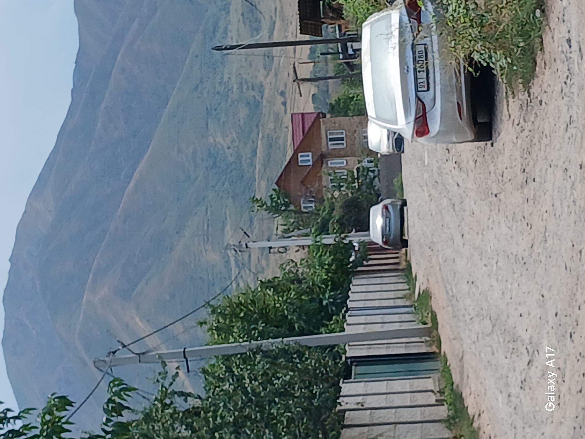 A dusty road winds through the village of Arashan, Kyrgyzstan, with a backdrop of rolling, arid mountains. On the left side of the road, a concrete fence with intermittent metal posts delineates a property line. Trees and foliage grow on either side of the fence. Several cars are parked or driving on the road. In the foreground, a white sedan is visible from the rear, its license plate reading "16080". Further down the road, another white sedan is parked. To the right of the road, a two-story house with a red roof and stone facade is visible, surrounded by trees. The overall scene depicts a quiet, rural setting in the mountains.