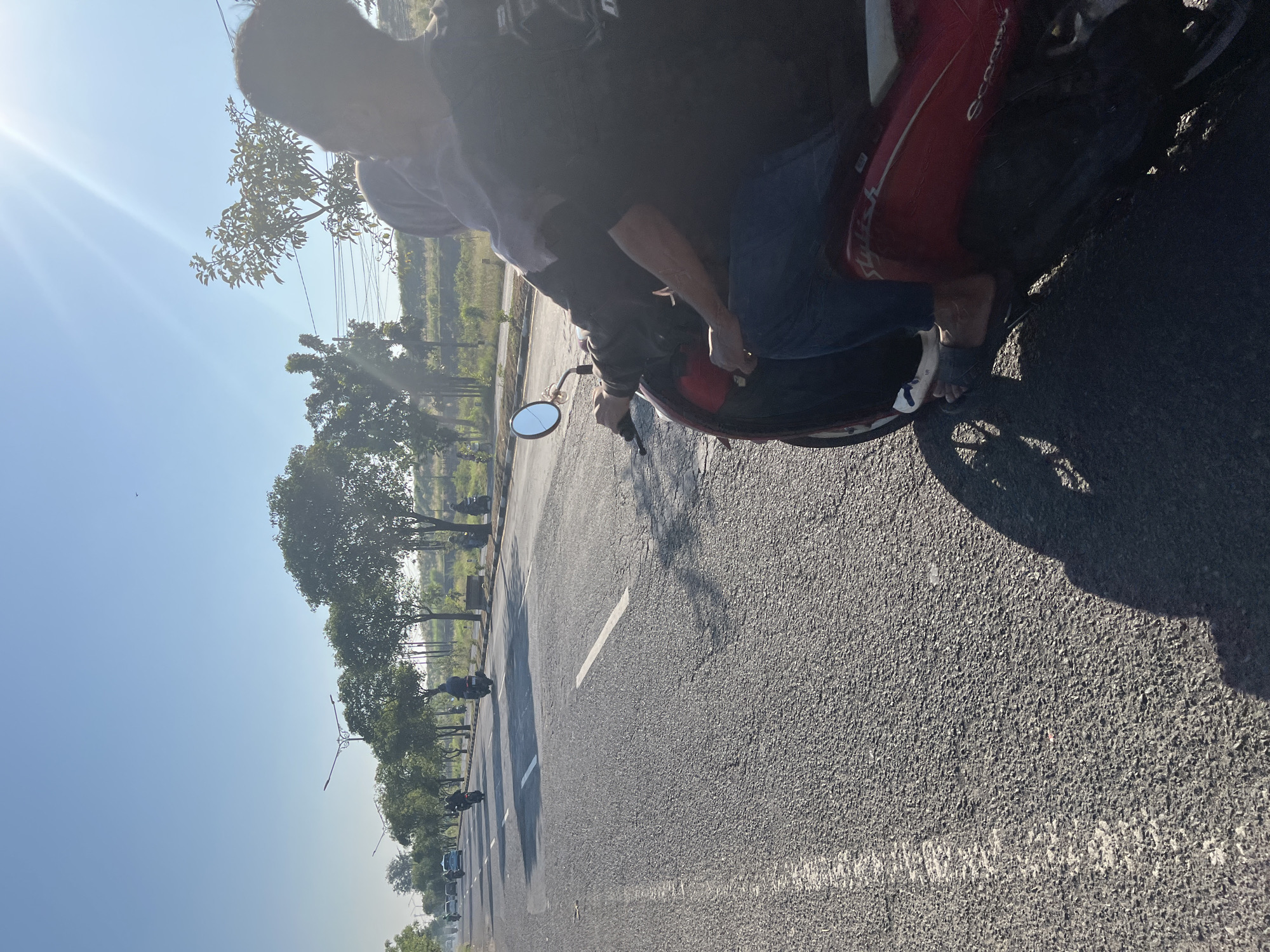 In Ketapang, Indonesia, a person is riding on the back of a red scooter on a paved road with white lane markings. The scooter is moving from right to left in the frame. The rider is wearing a black hoodie and blue jeans, and the person in front is wearing a hijab. The road is lined with trees and streetlights. The sun is shining brightly, casting shadows on the road. Other vehicles, including motorcycles and cars, are visible in the background on the road. The sky is clear and blue.