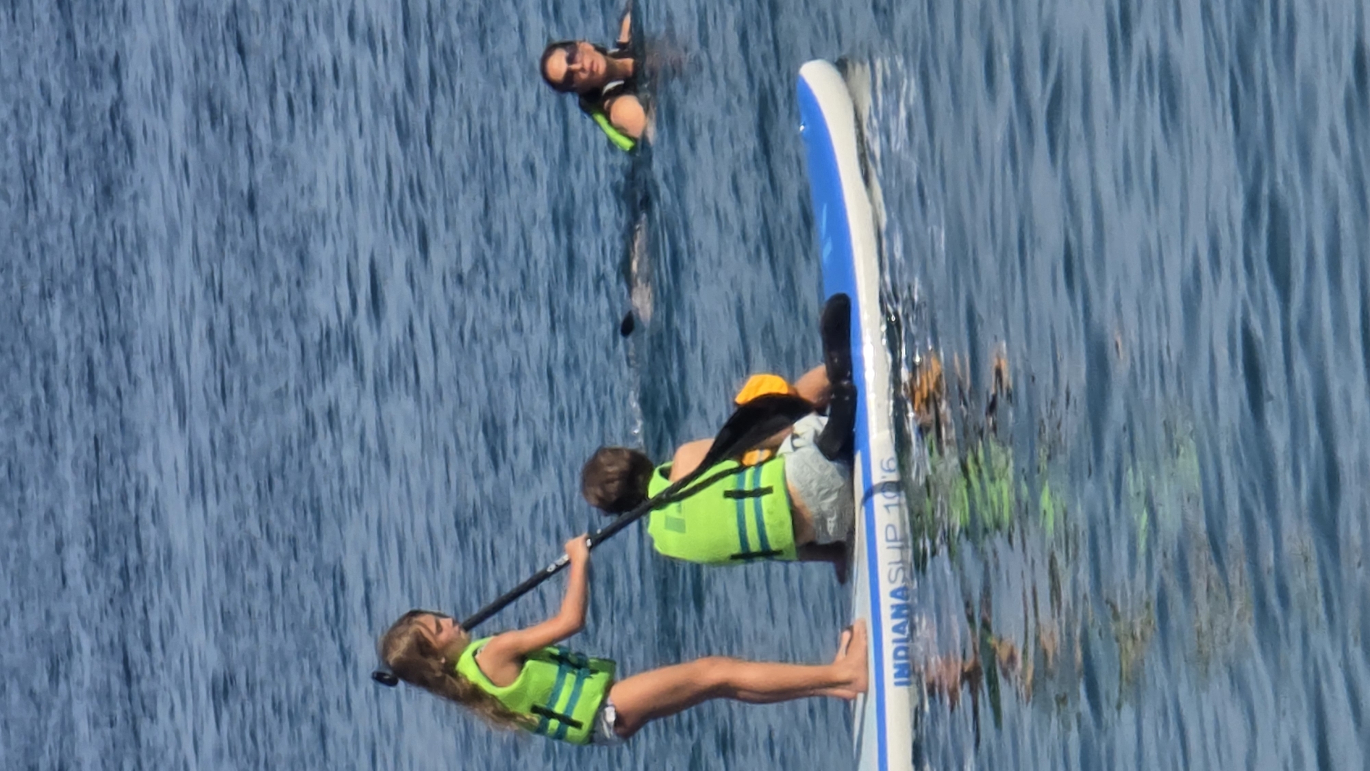 In this media file, three people are engaged in water activities on a sunny day. Two individuals, appearing to be children, are on a paddleboard. One is seated and holding a paddle, while the other is lying down, also wearing a bright green life vest. The paddleboard is blue and white with the text "INDIANASUP 10'6" printed on its side. A third person, an adult, is in the water nearby, also wearing a green life vest. The water is a deep blue with gentle ripples, suggesting calm conditions. The overall scene appears to be a recreational outing, possibly at a lake or the ocean, under bright daylight.