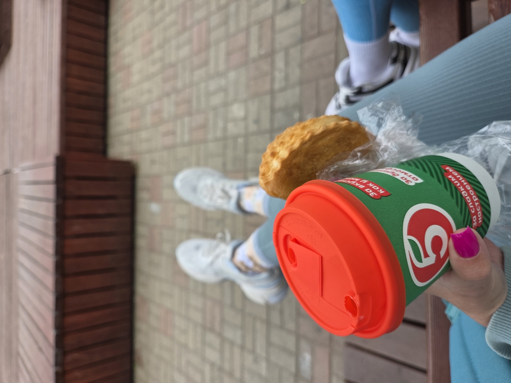 A top-down shot shows a person seated on a bench. They are wearing light blue leggings, white socks, and white sneakers with black accents. The bench is situated on a brick pavement with some wooden structures nearby. The person is holding a green coffee cup with a bright red lid, next to a packaged pastry. Their fingernails are painted hot pink. The cup has Russian text and a prominent number "5" logo on it. Given the logo and Russian text, it is likely the photo was taken in Russia. The lack of snow suggests it is not winter. The location may be Birobidzhan, Russia.