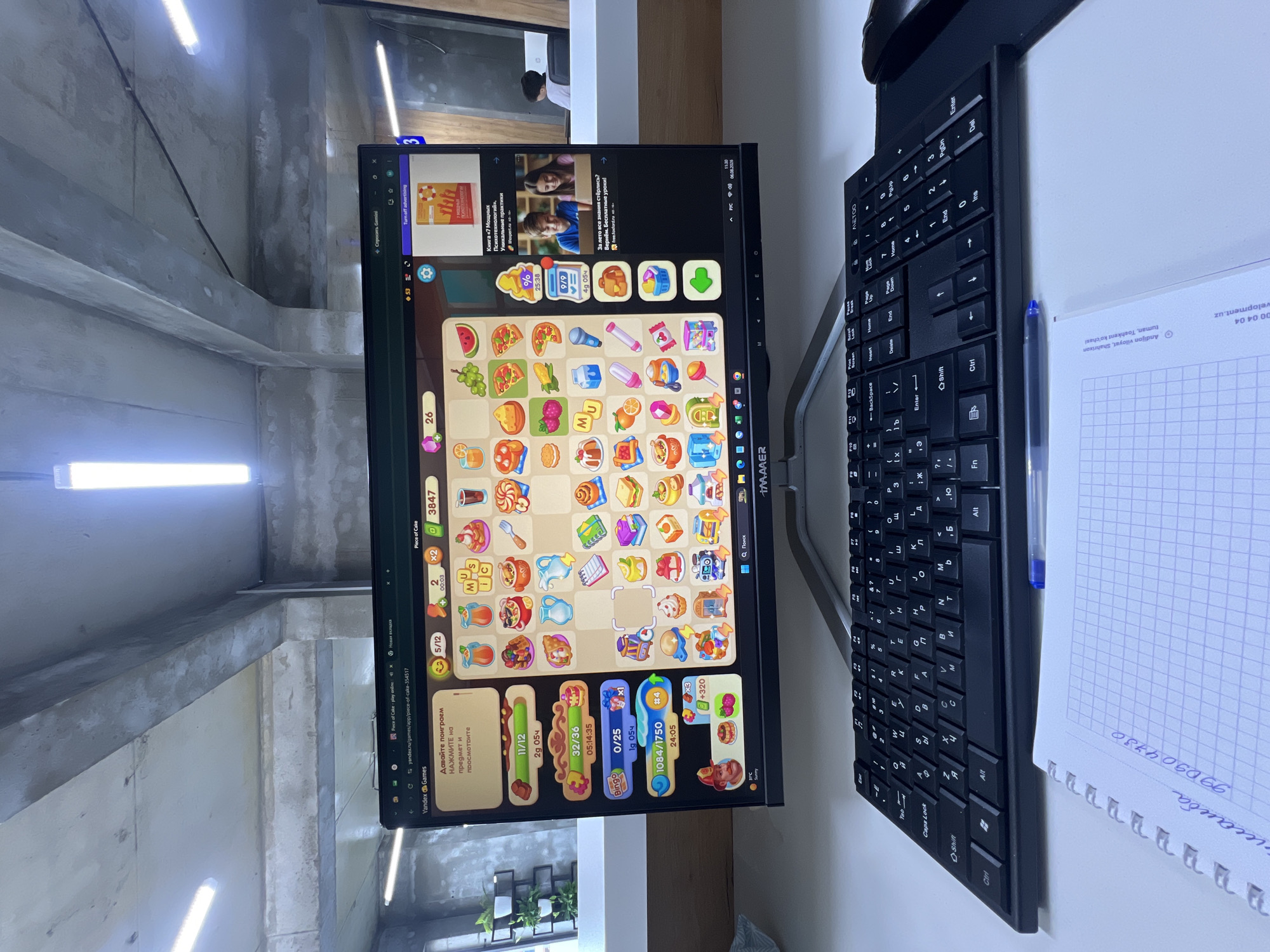 An indoor shot captures a computer monitor displaying a game called "Piece of Cake" on Yandex Games, situated on a desk in what appears to be an office environment. The monitor is positioned in front of a black keyboard and a notebook. The game interface shows various food-themed items and progress bars. To the right of the game, an advertisement for a book and a course is visible. In the background, there are concrete pillars and fluorescent lighting, suggesting an industrial or modern office space. The notebook in the foreground has handwritten text, including "Andijon viloyati Shahrixan" and a phone number, indicating a connection to Shahrixon, Uzbekistan.