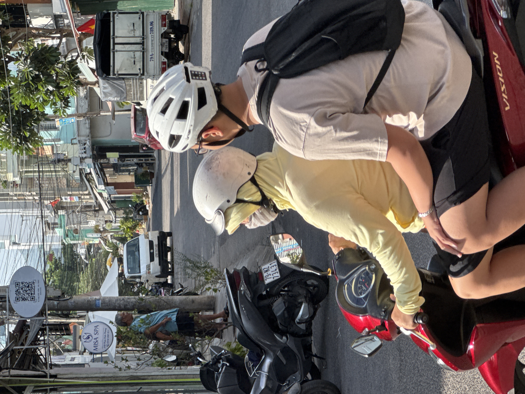 The image depicts a street scene in Nha Trang, Vietnam, possibly on a sunny day. In the foreground, two people are riding on a red scooter. The person in the front, wearing a yellow long-sleeved shirt and a beige helmet, is partially obscured. The person in the back, wearing a light-colored t-shirt and a white helmet, is facing away from the camera and has a black backpack. They are both wearing helmets, suggesting a local mode of transportation.
The scooter is parked on a paved street. To the left of the scooter, there is a parked dark-colored motorcycle. In the background, a white van is visible on the street, along with several buildings of varying heights and colors. One building has a sign for "MISA SPA." Power lines and street signs are also present, indicating an urban environment. The overall scene suggests a typical street in a Vietnamese city, with a focus on the everyday activity of using scooters for transportation.