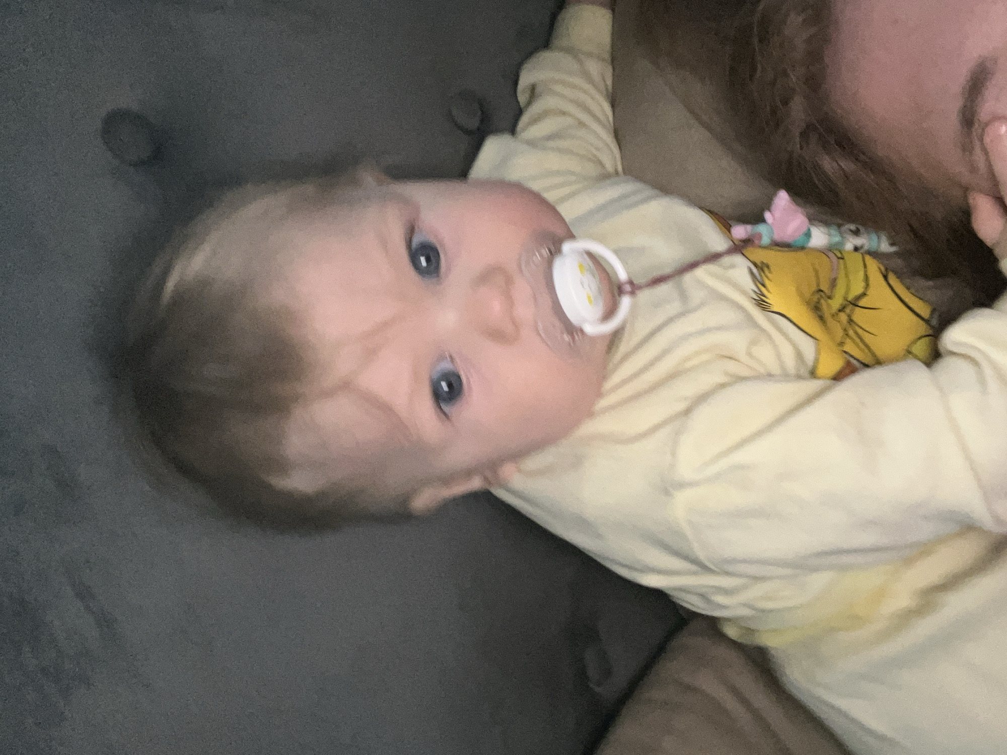 A baby with blond hair and blue eyes is lying on a gray sofa, facing upwards. The baby is wearing a yellow long-sleeved shirt and has a pacifier in their mouth with a white circular guard. A brown cord is attached to the pacifier. Part of a person with red hair is visible to the right of the baby, and there is a yellow blanket with an image of a cartoon character. The location in the city of Vesyoloye, Russia, cannot be determined from the image.