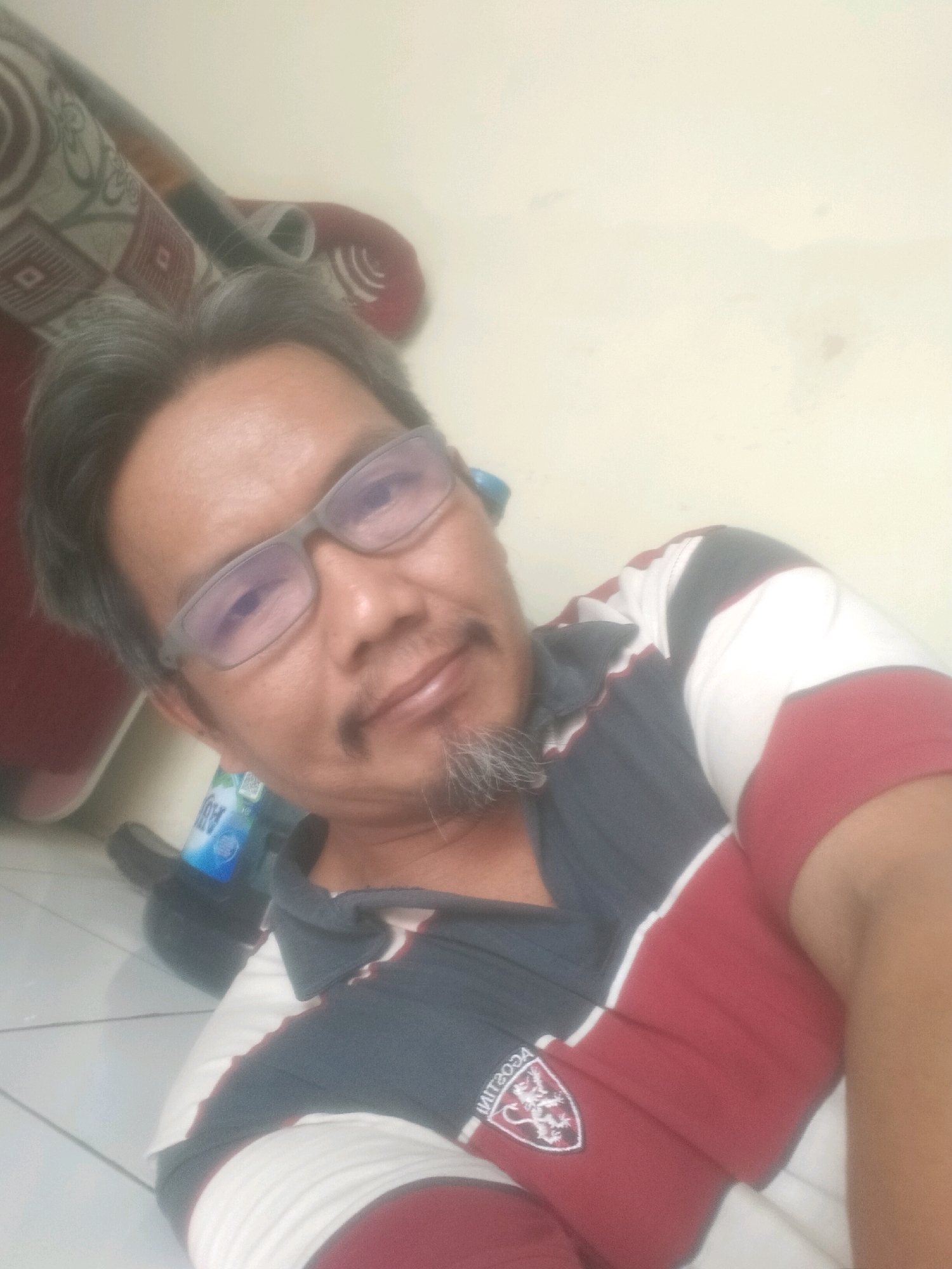 A man with graying hair and a goatee is lying on a tiled floor, leaning against a light-colored wall. He is wearing a collared shirt with navy blue, white, and maroon stripes. He has on glasses. To his left, a bottle of Aqua water is visible. Behind him, a sofa with a patterned fabric can be seen. The image appears to be a selfie taken indoors, likely in a residential setting in Suka Sari, Indonesia.