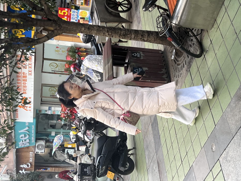 This image depicts a daytime street scene on a sidewalk in Jiangjin, Chongqing Municipality, China. The primary subject is an adult female, approximately 30-40 years old, with dark hair pulled back, wearing clear-framed glasses, a long pale pink or beige puffer coat, light blue wide-leg pants, and white athletic shoes. She walks from left to right across the tiled sidewalk, looking slightly downward and to her left. She carries a small brown patterned shoulder bag with a reddish-pink strap, appearing to be a Louis Vuitton monogram design, on her left side, and holds a dark-colored smartphone in her right hand.
The sidewalk features green square tiles interspersed with darker grey sections. To the right, a tree with thin branches stands, and a brown wooden double trash receptacle with recycling symbols is positioned near its base. Further right, a black bicycle is partially visible, leaning against a metal barrier. In the background, numerous parked scooters and electric motorcycles, some covered, line the street. One black scooter displays a "渝A 622VM" license plate. Storefronts with Chinese signage are visible behind the parked vehicles. Notable signs include "千色 美发工作室" (Qian Se Hair Salon Studio), "洗e人" (Wash-e-people), "李时珍中医医疗" (Li Shizhen Traditional Chinese Medicine Medical clinic), and a blue sign advertising "助听器" (Hearing aids) with a "2.5" sales indicator. An air conditioning unit is mounted on the wall of one shop. On the far left, another individual in a red coat is partially visible in the distance. An old-fashioned cart with large wooden wheels is visible on the far right of the background.