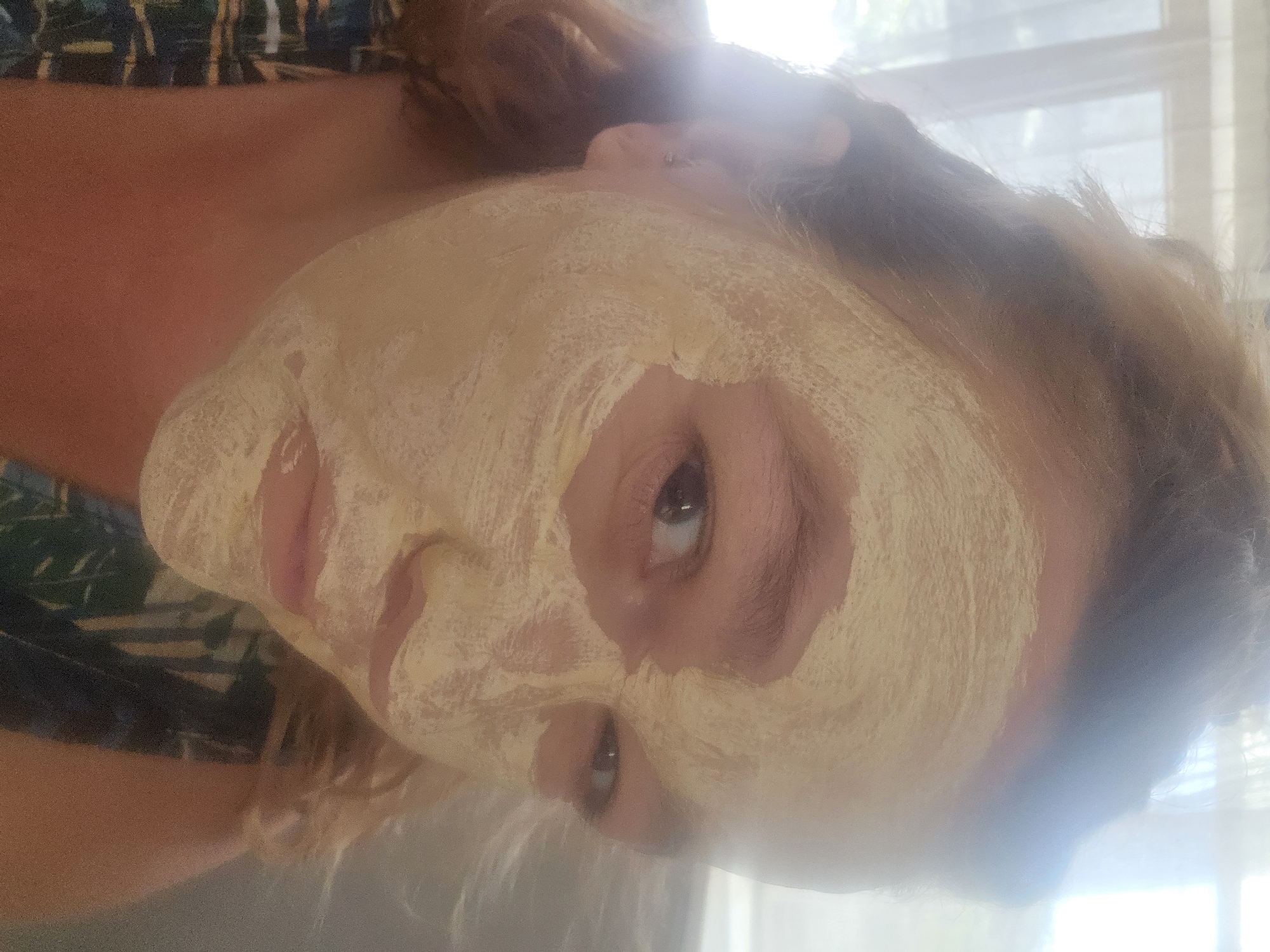 A close-up, medium shot captures a woman with her face covered in a light yellow facial mask, looking directly at the camera. She has medium-length, wavy blonde hair. The woman is wearing a patterned top, possibly with dark blue and green floral motifs. Her left ear is visible, adorned with a small earring. The lighting in the image is bright and somewhat overexposed, with a blurry background suggesting an indoor setting with a window providing natural light. The facial mask covers most of her face, obscuring her nose and upper cheeks, but her eyes and lips are visible. One eye is looking directly at the camera, while the other is slightly averted. The overall impression is of a person engaged in a skincare routine. The location is not determinable from the image.