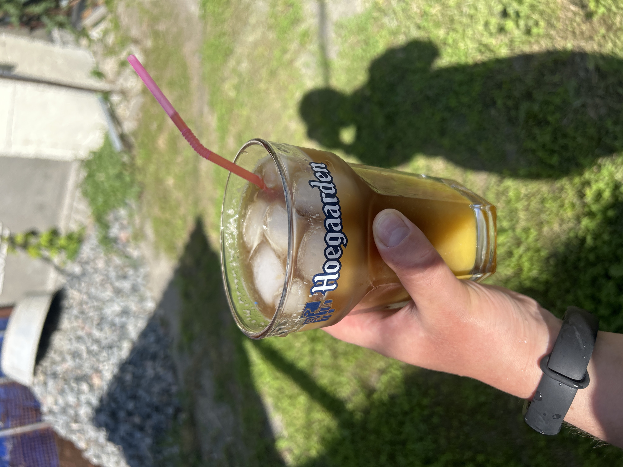 A hand holds a glass of iced beverage with a pink straw. The glass has a blue and white logo that reads "Hoegaarden". The beverage appears to be amber in color. The background is out of focus, showing green grass, a pile of gravel, and a building. The scene is lit by sunlight, casting shadows. There is no visible indication of the location being the city of Bila Tserkva, Ukraine.