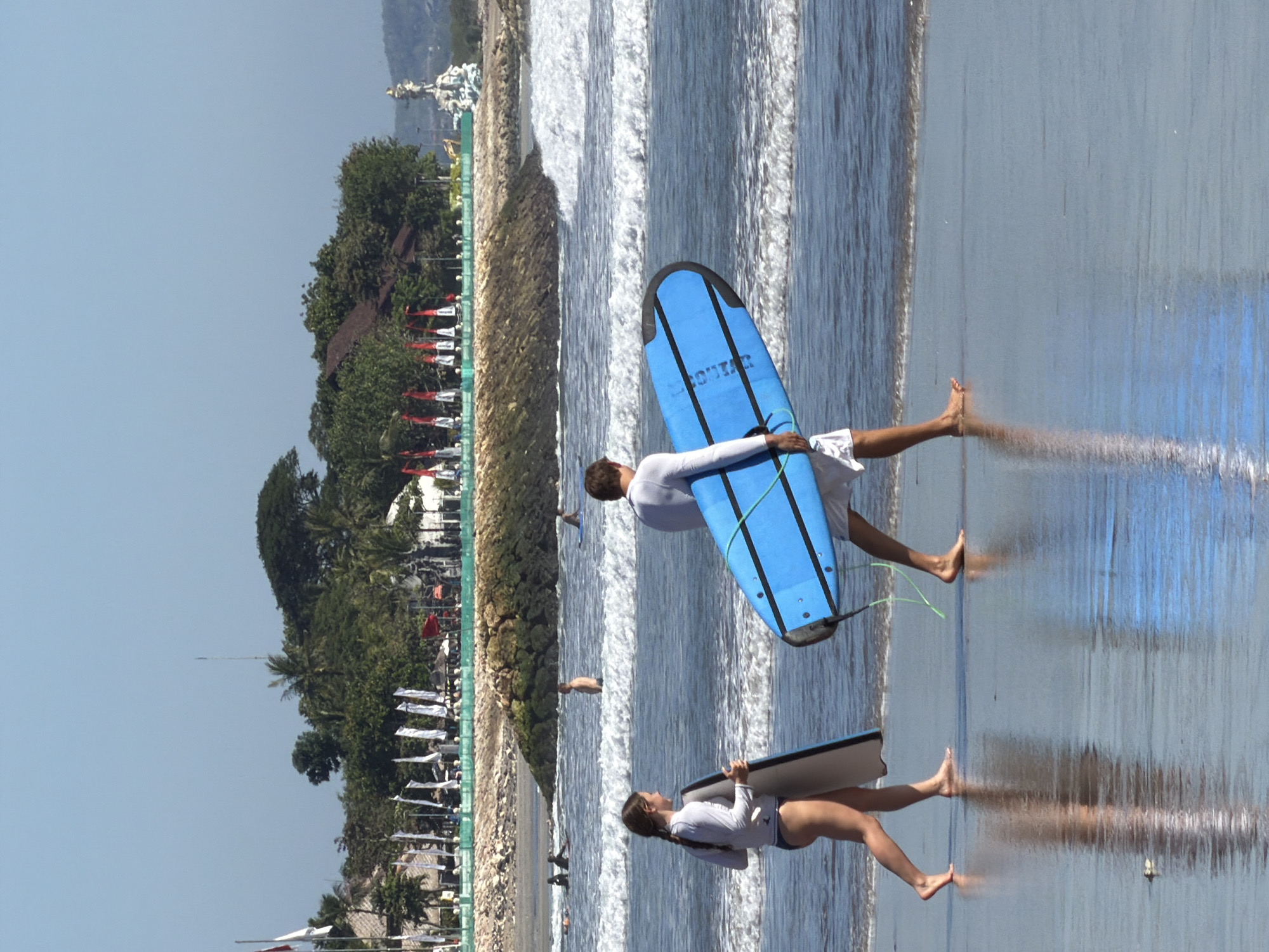 Two people, a woman and a man, walk along the wet sand of Kuta Beach in Bali, Indonesia, carrying surfboards. The woman, on the left, wears a white long-sleeved rash guard and dark bikini bottoms. She carries a dark surfboard tucked under her left arm. The man, on the right, wears a white long-sleeved shirt and white shorts. He carries a bright blue surfboard. Both individuals appear to be walking towards the right side of the frame. The ocean, with moderate waves, is to their right. In the background, a rock jetty extends into the water, and beyond that, lush green trees and buildings are visible. A statue and flags are also present on the coastline. The sky is clear and blue.
