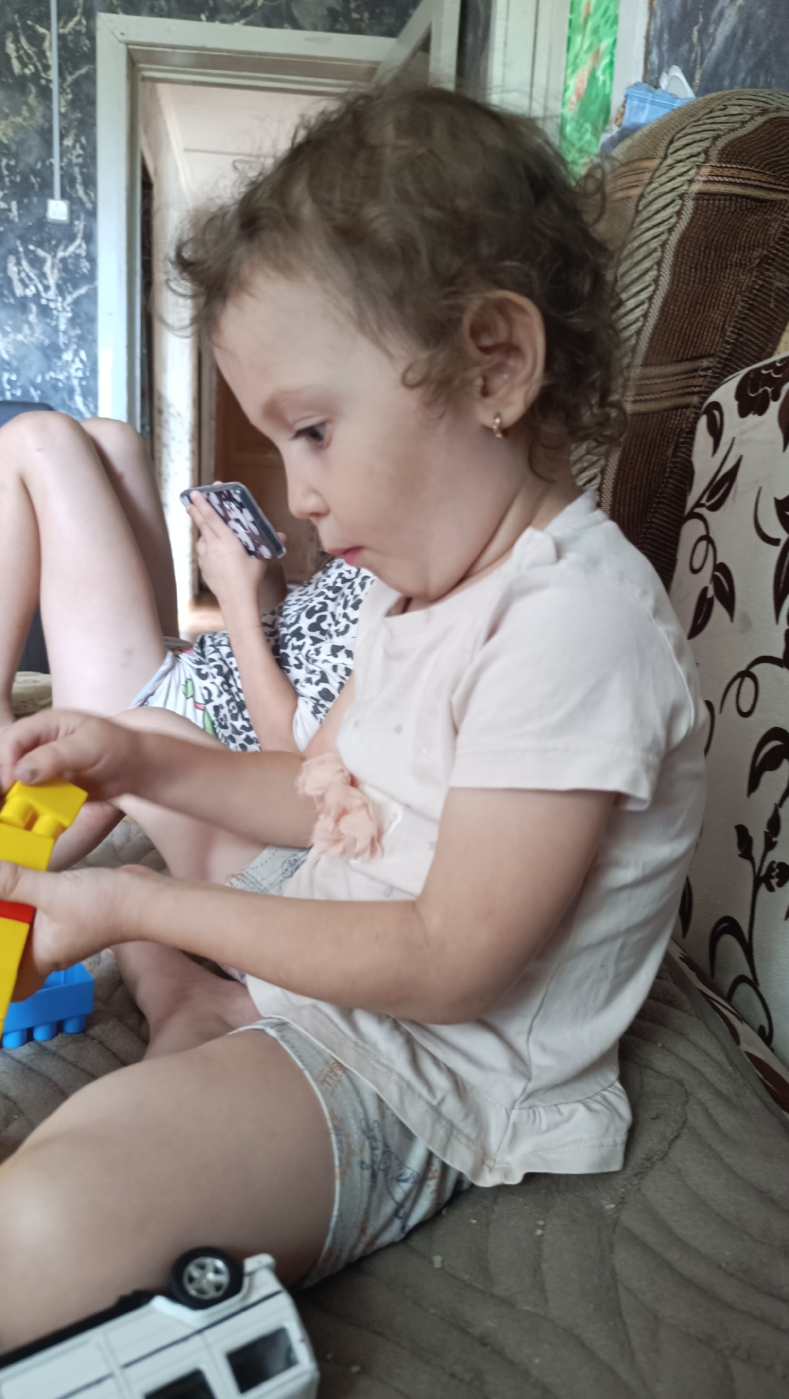 A young child with curly brown hair is sitting on a couch in a room. The child is wearing a light pink t-shirt and light-colored shorts with a patterned design. The child is holding yellow and blue toy blocks in their hands. To the left of the child, another person's legs are visible, with a phone in their hand. In the foreground, a white toy bus is on the couch. The room appears to be a living space, with a patterned couch and what looks like a doorway in the background. The overall impression is of a casual, domestic scene. The image does not provide enough information to determine the location within the city of Karsha, Russia.