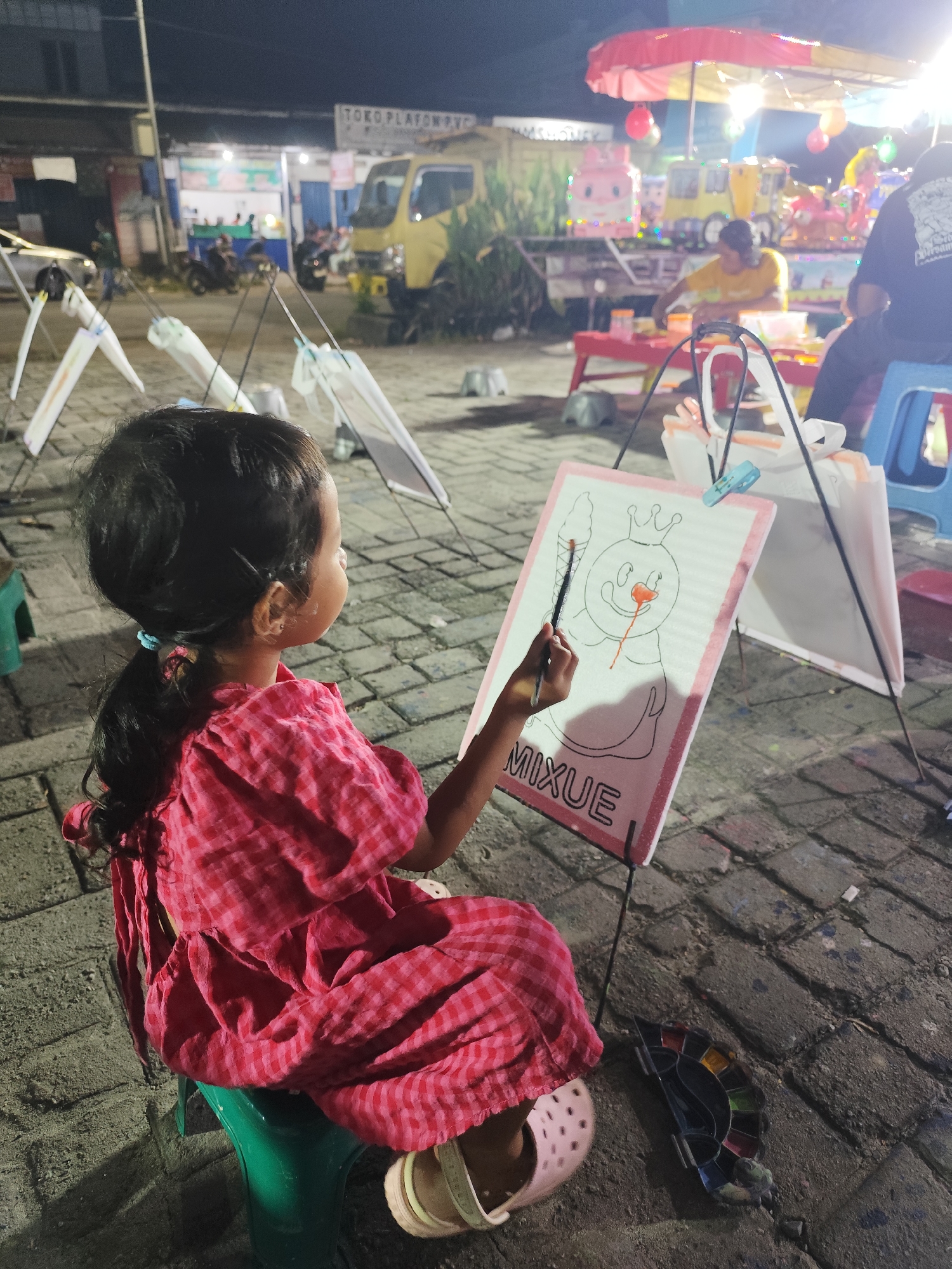 A young girl in a red checkered dress sits on a green stool, facing away from the camera, engrossed in painting. She holds a paintbrush in her right hand and is adding color to a drawing on a white board propped up on an easel. The drawing depicts a character with a crown and a nose that appears to be colored orange. Below the drawing, the word "MIXUE" is visible in large, block letters. A palette of watercolors is on the ground next to her.
The scene appears to be outdoors at night, possibly at a market or fair. In the background, there are stalls with lights and people, and a yellow truck is visible. Other easels with white boards are set up in the foreground, suggesting an art activity area. The ground is paved with cobblestones. The overall mood seems peaceful and focused, with the young artist concentrating on her creative endeavor.