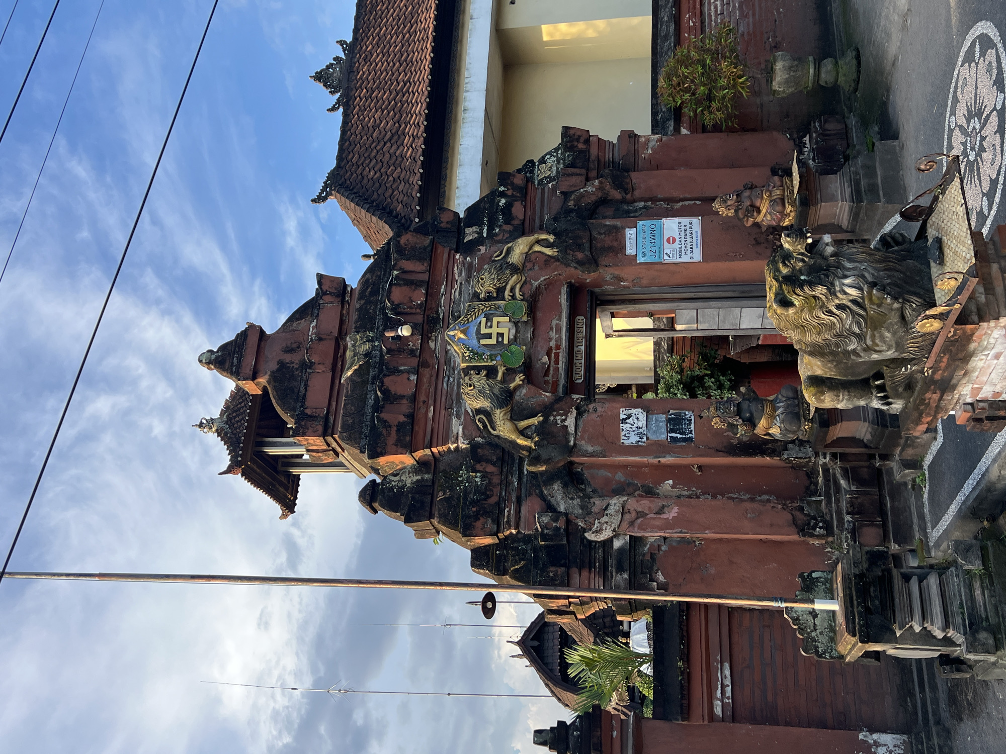 The image depicts an ornate entrance gate, likely to a building or complex, in Singapadu, Indonesia. The structure is predominantly red brick with intricate carvings, including golden lion-like figures and a square emblem with a swastika. The gate is weathered and partially covered in moss or lichen. There is a sign posted on the gate with the text "TUOK MD TRESINA" and "ROMAH SEHAT RAP STASIUN KRAP JZ14WNQ MOBIL DAN MOTOR MOHON PARKIR DI JABA (LUAR) PURI". A stone planter is visible, along with a tile mosaic on the ground, and an external lighting fixture. Above the gate are building rooflines. The sky is visible in the background with a light cloud cover, and electrical wires running across the top.