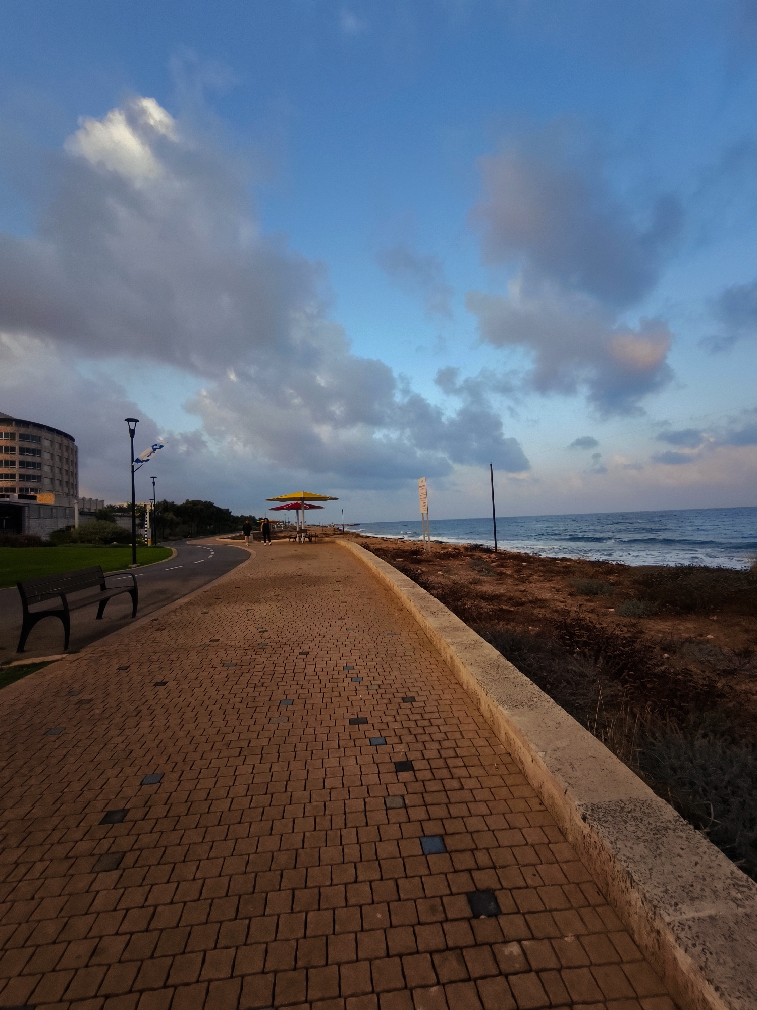 This is a vertical, eye-level shot of a paved promenade and the sea in Nahariyya, Israel, likely taken at dusk. The promenade is paved with interlocking rectangular bricks of a reddish-brown hue. A dark wooden bench with black metal legs sits to the left of the promenade. Further along the promenade, two individuals, one walking and one standing, are near a colorful umbrella structure with red and yellow panels, accompanied by picnic tables. Beyond the promenade, a strip of dry, brown vegetation separates it from the calm, blue sea, where gentle waves are breaking on a sandy shore. In the background, a multi-story, light-colored building with numerous windows is visible, adjacent to a well-maintained green lawn and trees. The sky above is a vibrant blue, dotted with scattered, wispy clouds, some of which are tinged with pink and orange, suggesting the last light of sunset. The overall atmosphere is serene and coastal.