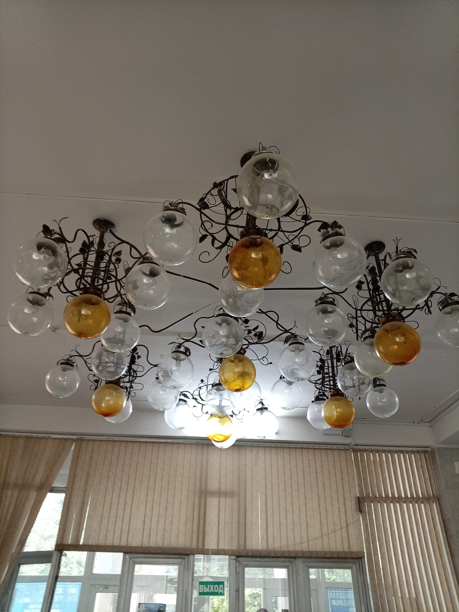 The image shows a chandelier with multiple glass globes hanging from a white ceiling. Some of the globes are clear, while others are amber-colored. The chandelier has an ornate metal frame. Below the chandelier, there are vertical blinds covering windows, and through the windows, it appears to be daytime with some greenery visible. A green sign with Russian text, "ВЫХОД" (Exit), is visible on one of the windows. The setting appears to be an indoor public space. This scene is located in the city of Zheleznovodsk, Russia.