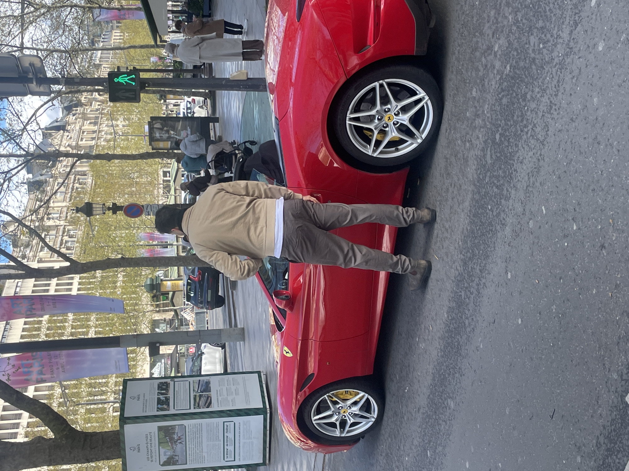 A forensic, matter-of-fact description of the scene follows:
This image captures an urban street scene during daylight hours on the Champs-Élysées in Paris, France. The location is confirmed by a green information kiosk on the left displaying text about "PARIS" and "LES CHAMPS-ÉLYSÉES" renovation work, and banners advertising "ART PARIS GRAND PALAIS".
In the foreground, a bright red Ferrari convertible, appearing to be a California T or Portofino model, is positioned on the dark grey asphalt street. The vehicle's convertible top is down, and it features silver multi-spoke wheels with yellow brake calipers and Ferrari center caps.
Standing next to the driver's side of the Ferrari, viewed from behind, is a male individual. He is dressed in a beige jacket, a white t-shirt visible beneath, dark grey trousers, and dark brown boots. His posture suggests he is either standing beside the vehicle or interacting with it.
The street surface shows patches of wetness. On the paved sidewalk to the right, several pedestrians are visible. One individual in a long, light-colored coat stands near a pole with a green "walk" signal. Further left, another person pushes a baby stroller. In the background, classic Parisian Haussmannian-style buildings with multiple stories line the street. Bare deciduous trees with nascent green foliage are interspersed along the thoroughfare, framing the upper portion of the scene against a partially cloudy blue sky. Other vehicles, including a dark SUV and a white car, are visible in the background. Street furniture includes various poles, a dark lamppost, and a "No Parking" sign.