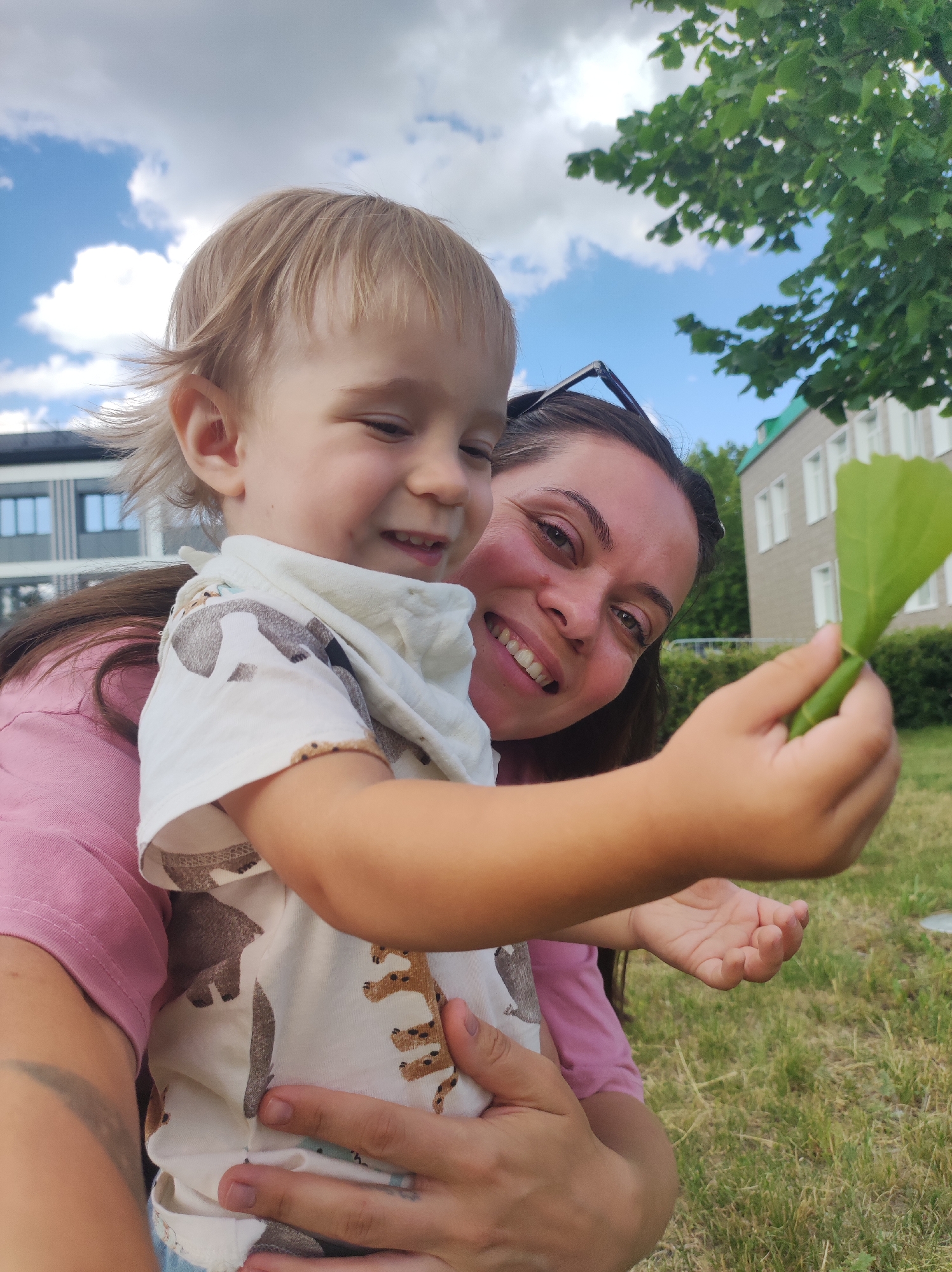 In this candid, eye-level outdoor portrait, a woman and a young child are the central subjects, smiling at the camera. The woman, positioned behind and to the right of the child, has dark hair and is wearing a pink top. She is holding the child, who has light blonde hair and is wearing a white t-shirt with a brown animal print. The child is looking down and to the left, with a joyful expression, while the woman is looking directly at the camera with a wide smile.
The woman is holding a bright green leaf in her right hand, extending it towards the camera, as if showing it to the viewer. The child is wearing a light-colored cloth around their neck.
The setting appears to be a park or outdoor recreational area. Green grass covers the foreground, and in the background, there are trees with lush green leaves, a building with a light-colored facade and multiple windows, and a bright blue sky with scattered white clouds. The lighting suggests it is daytime, likely a clear and pleasant day. The overall mood is one of happiness and connection. There is no visible text in the image.
