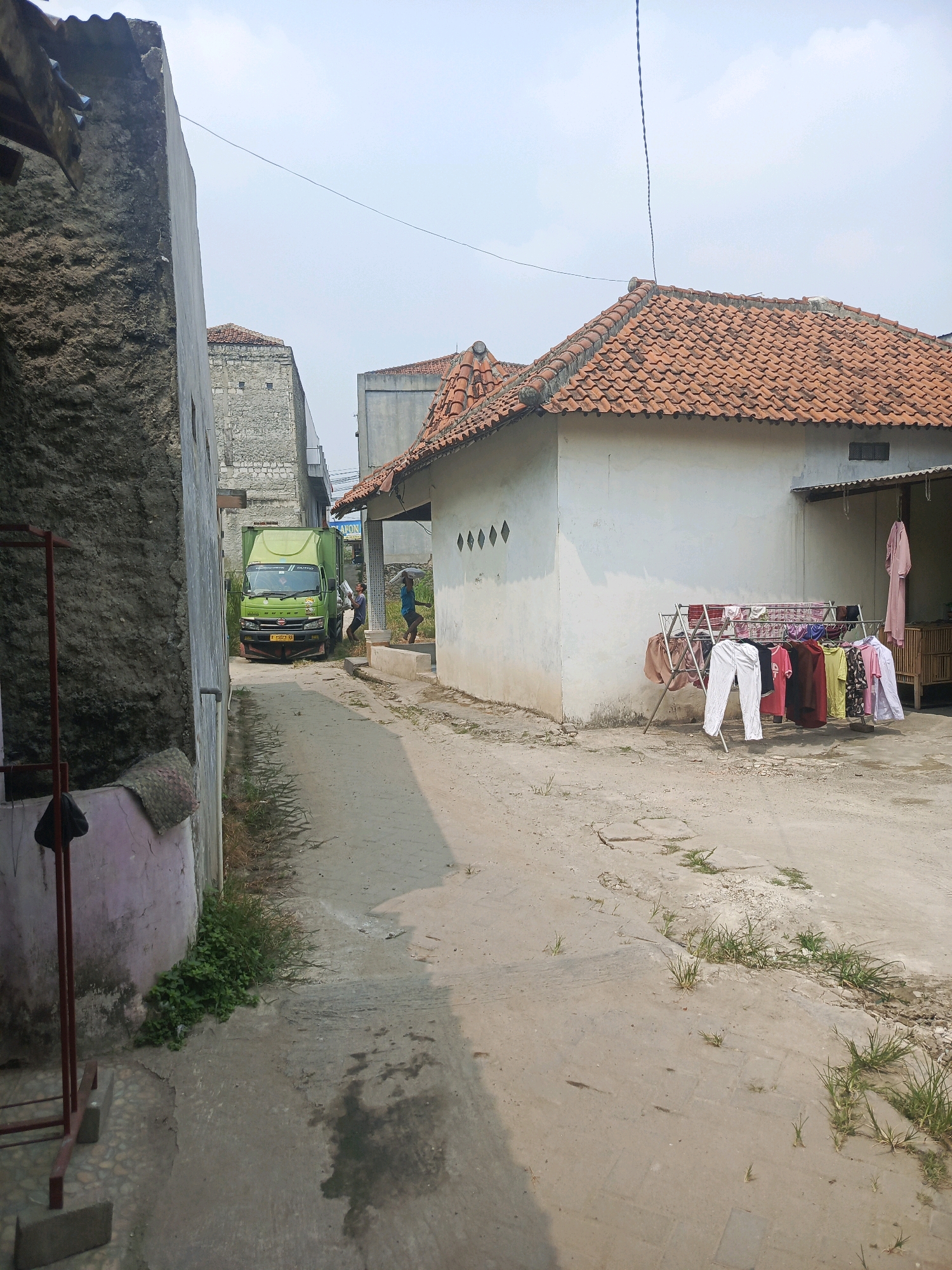 In the city of Balaraja, Indonesia, an alleyway is depicted with a concrete path that shows some cracking and weeds growing through it. On the left, a tall, rough-textured gray wall runs along the side of the alley. To the right, a white house with an orange tiled roof is visible. In front of the house, a metal drying rack is filled with various items of clothing, including white pants, colored shirts, and a pink garment hanging from a hook on the house wall. Further down the alley, a green truck is parked, with two men standing near it. One man is on the driver's side, and another is on the passenger side, appearing to interact with the truck. In the distance, a tall, stone-like structure with square openings can be seen. The sky is overcast.