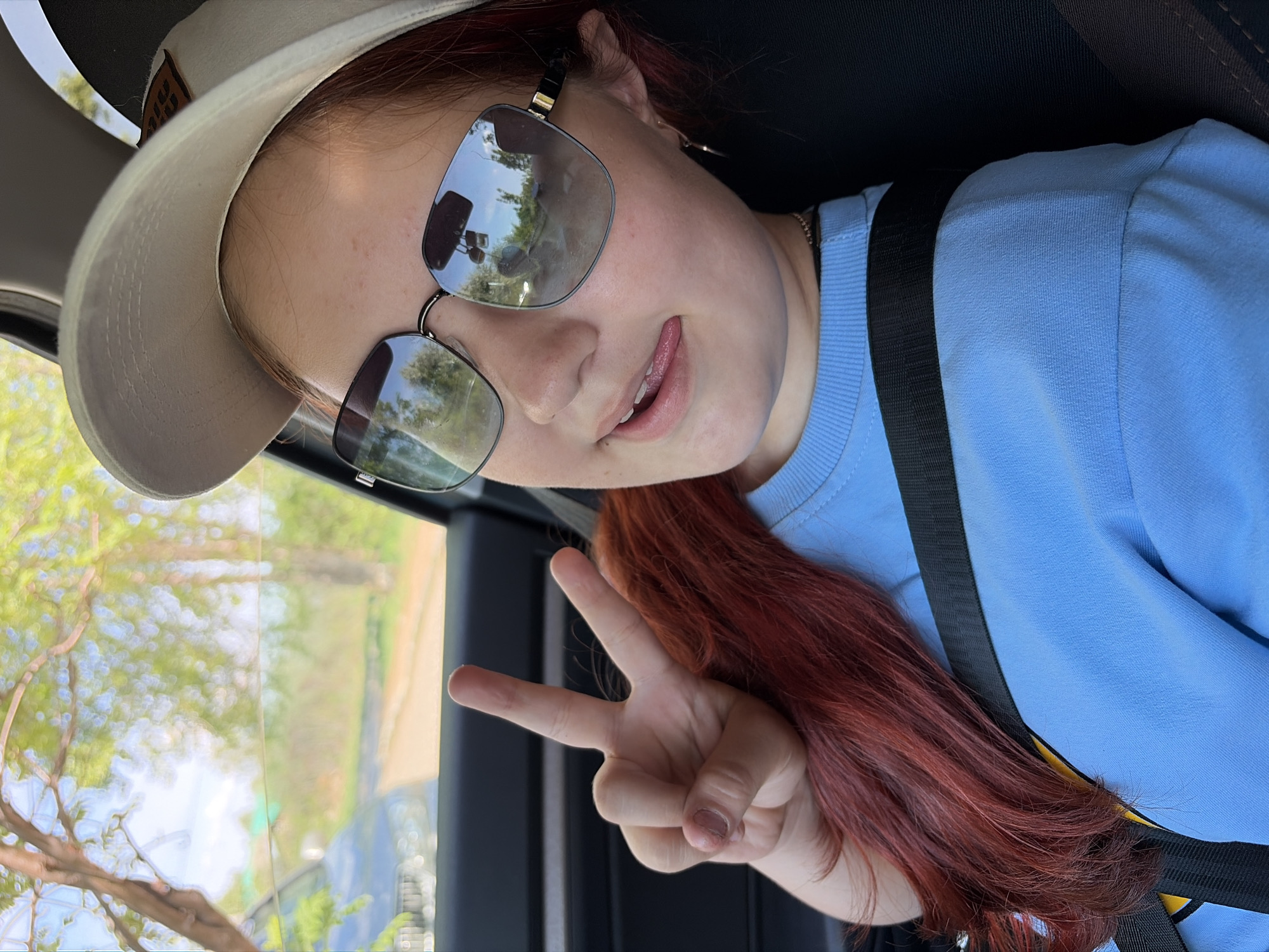 A young woman with red hair is sitting in a car, wearing a light blue long-sleeved shirt, a tan hat, and sunglasses. She is wearing a seatbelt and making a peace sign with her hand. The car is moving, and through the window, trees and a building can be seen. Based on the prompt, the location is in the city of Chaikovsky, Russia.