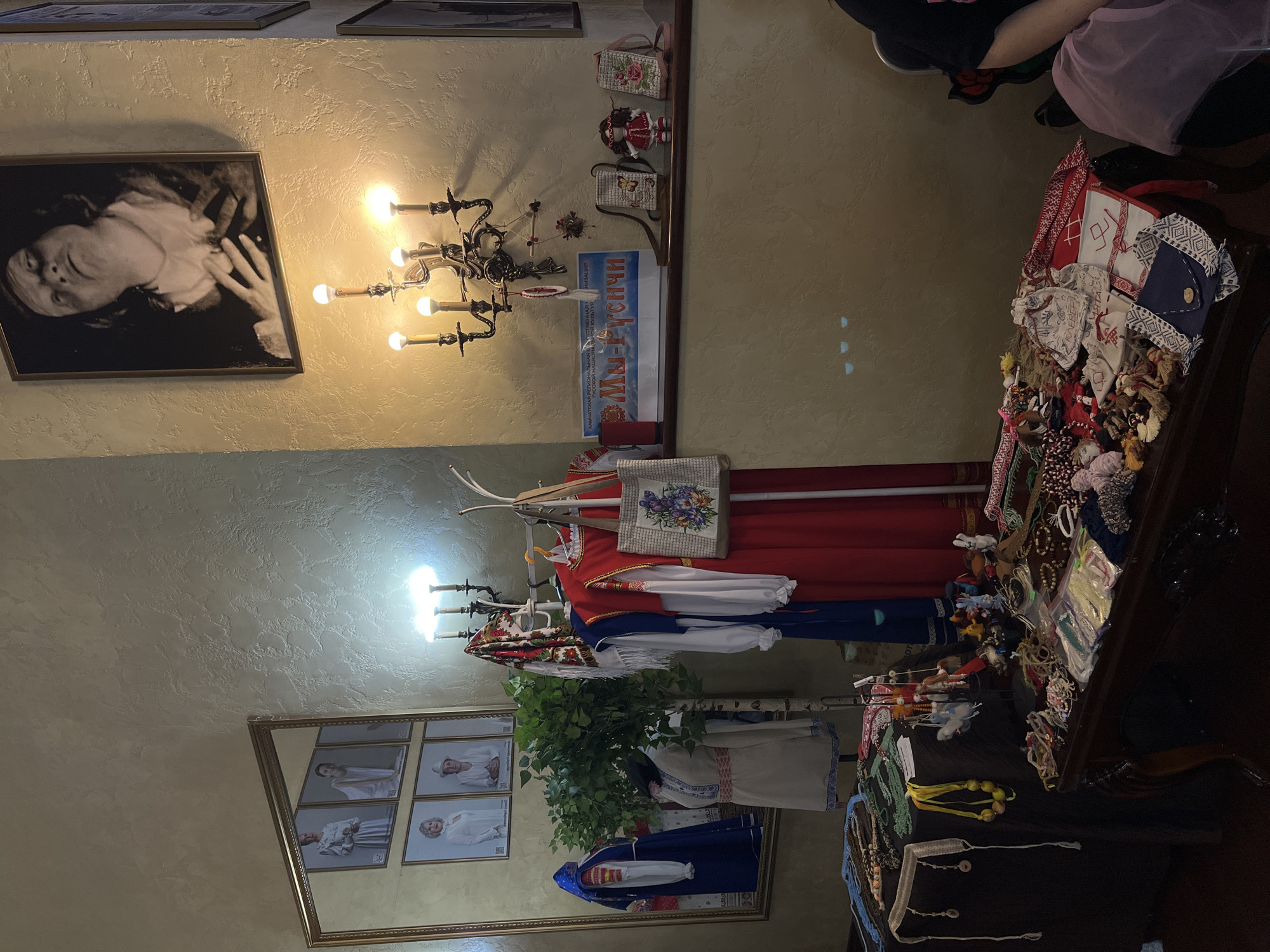 The image depicts a shop interior, likely in Petropavlovsk-Kamchatsky, Russia, given the "Мы-Русичи" (We are Rusichi) signage. The scene includes several cultural artifacts and clothing displayed for sale or exhibition. On the walls, there are framed pictures, including a black-and-white portrait and a collection of framed portraits possibly showcasing historical figures or residents. A decorative candelabra illuminates the upper part of the scene. Garments are hung on display racks and include a predominantly red dress with white and blue accents, along with other traditional Russian outfits. A handbag decorated with floral patterns is suspended from a hanger. A bushy plant adds a touch of nature to the interior. The foreground shows a table filled with crafts, jewelry, and other small decorative items. In the upper right corner, a person is visible, likely browsing the items on display.