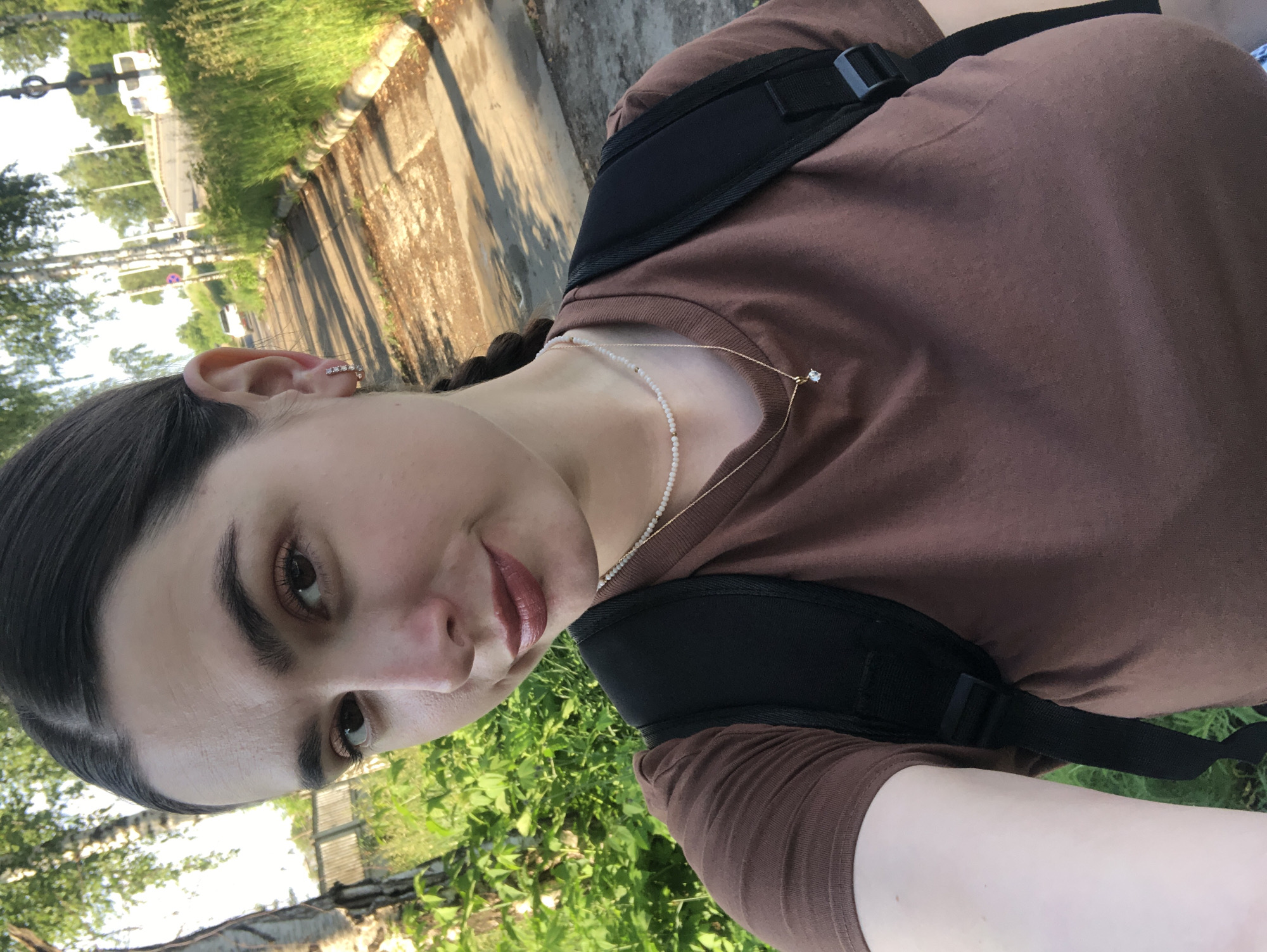 A young woman with dark hair pulled back in a braid, wearing a brown t-shirt, a pearl necklace, and a simple gold necklace with a diamond pendant, stands outdoors. She is wearing a backpack with black straps. The woman is on a paved path next to a grassy area with trees. In the background, there is a road with vehicles and more trees. The scene appears to be a park or a quiet street in the city of Chaikovsky, Russia.