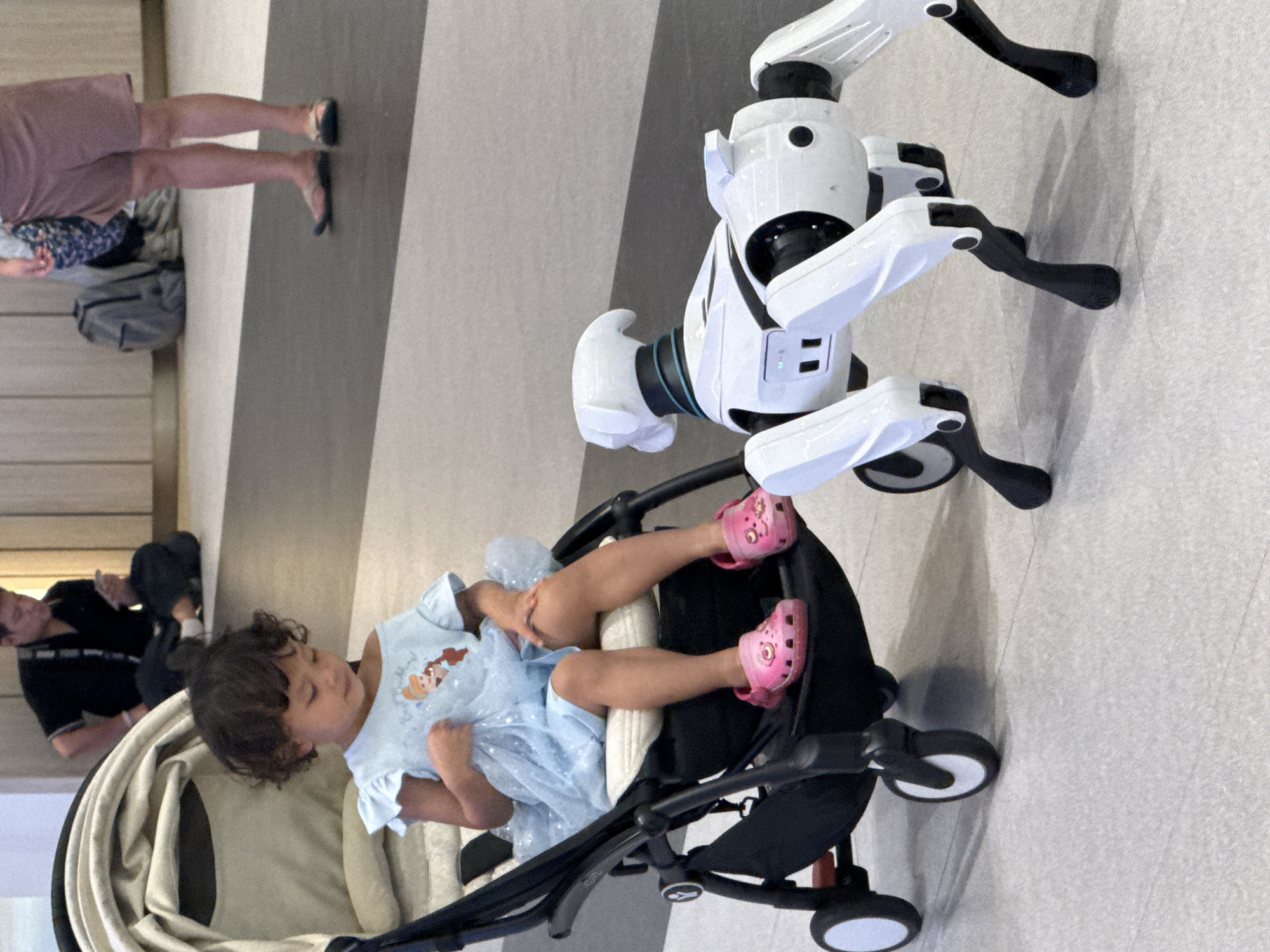 The image shows a young child, likely a toddler, sitting in a stroller. The child is wearing a light blue dress with what appears to be a Disney princess design and pink Crocs. The stroller is black with beige padding. Next to the stroller is a white robot with black accents, designed to look like a quadrupedal animal with articulated limbs and a distinctive head. The robot is standing on the floor, its pose suggesting it is interacting with or observing the child. In the background, parts of other people are visible, including legs in a pink skirt and sandals, and a person in a black shirt, suggesting a public setting such as a mall or an event. The floor appears to be tiled with a light grey pattern. The lighting suggests it is daytime. There is no visible text in the image. The scene is likely taking place indoors, possibly in Bangkok, Thailand, given the context. The overall mood appears casual and curious, with the robot's presence adding an element of novelty to the scene.