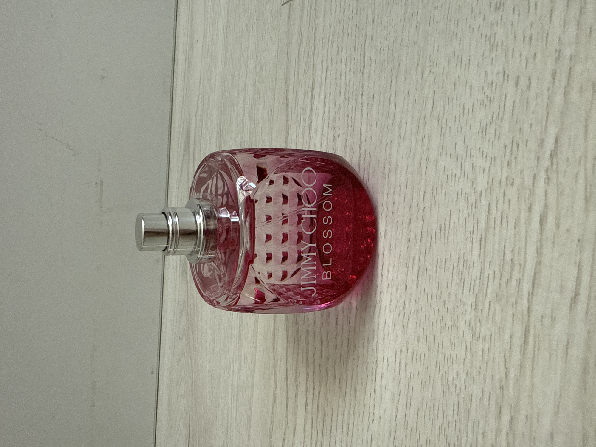 A bright pink Jimmy Choo Blossom perfume bottle stands on a light-colored wooden surface. The bottle has a faceted design and a silver cap. The background is a plain, light gray wall. The scene is indoors, with no indication of being in Burgas, Bulgaria.