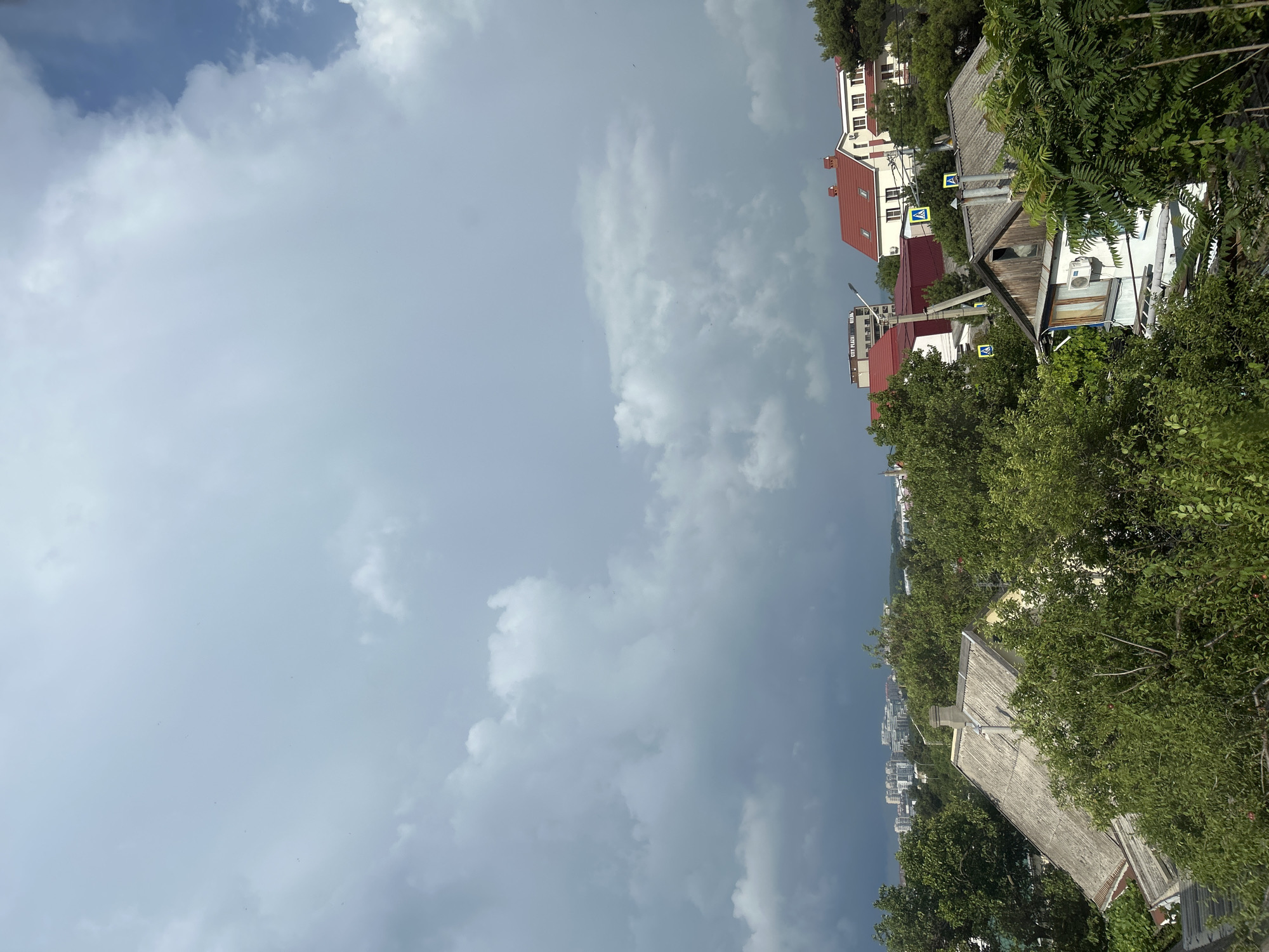 The image captures a view of rooftops and dense green foliage against a dramatic, cloudy sky over the city of Gelendzhik, Russia. In the foreground, several houses with tiled roofs are visible, interspersed with lush trees and bushes. One house in the mid-ground features a light-colored wall with a window and an air conditioning unit. Further back, a collection of apartment buildings and other structures can be seen, with a hint of the sea in the distance. The sky is filled with large, gray clouds, suggesting an overcast or impending storm. The overall scene conveys a sense of suburban residential life within a coastal city.